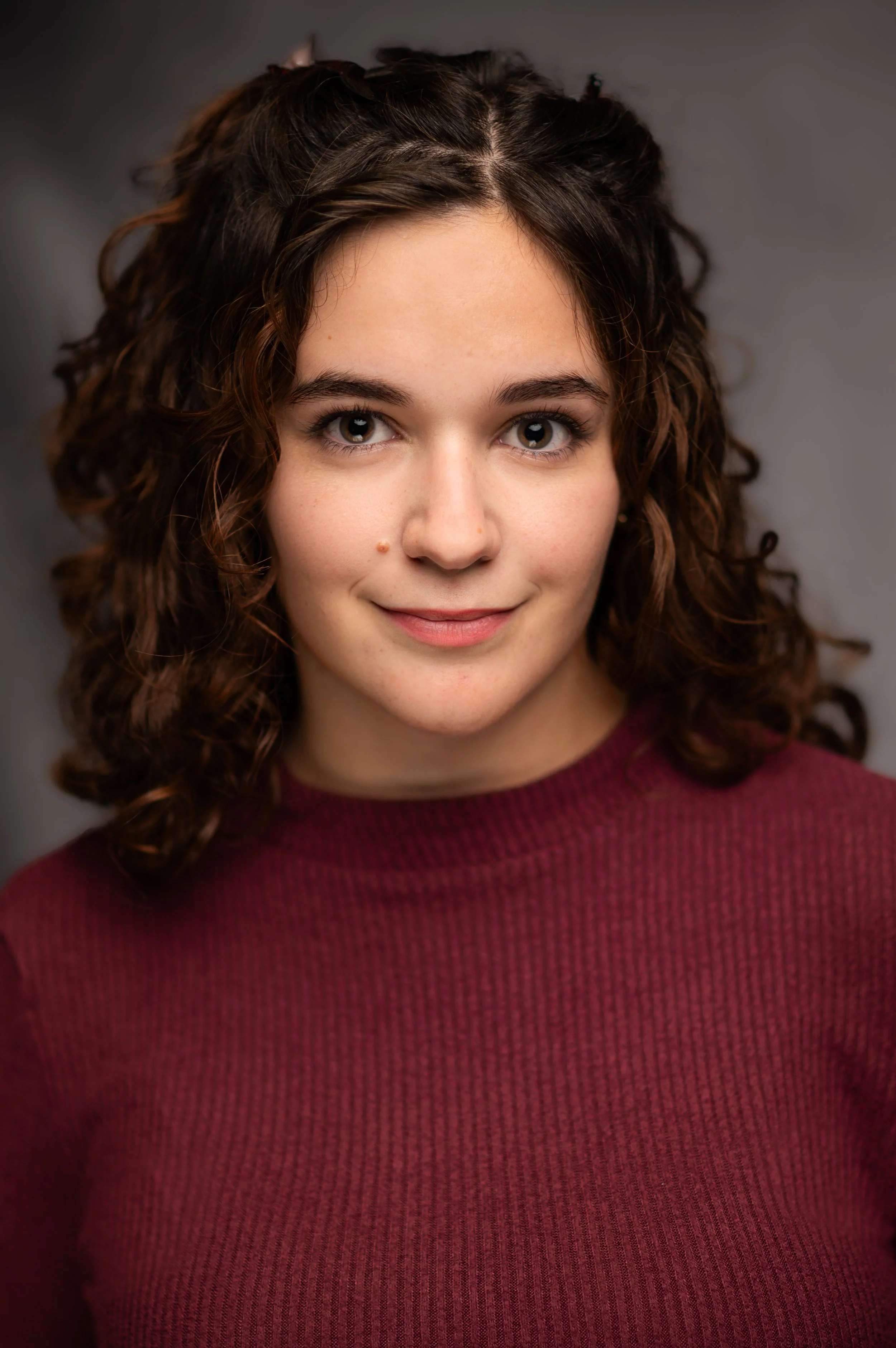 A close-up portrait of a young woman with curly brown hair, fair skin, and gray eyes, wearing a maroon sweater and smiling subtly against a gray background.