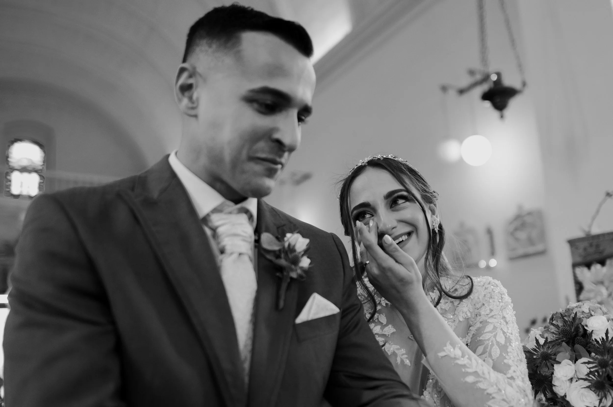 A black-and-white photo of a bride and groom at their wedding ceremony, smiling and looking at each other.