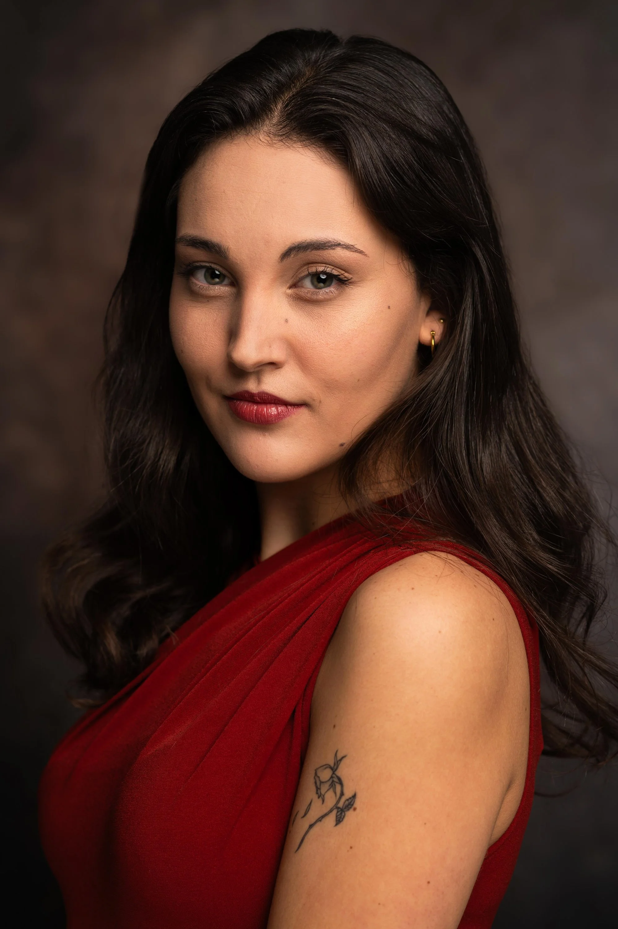 Close-up portrait of a woman with dark wavy hair, wearing a red dress, with light makeup, gold earrings, and a tattoo of a leafy branch on her upper arm. She is looking at the camera with a slight smile against a dark, blurred background.