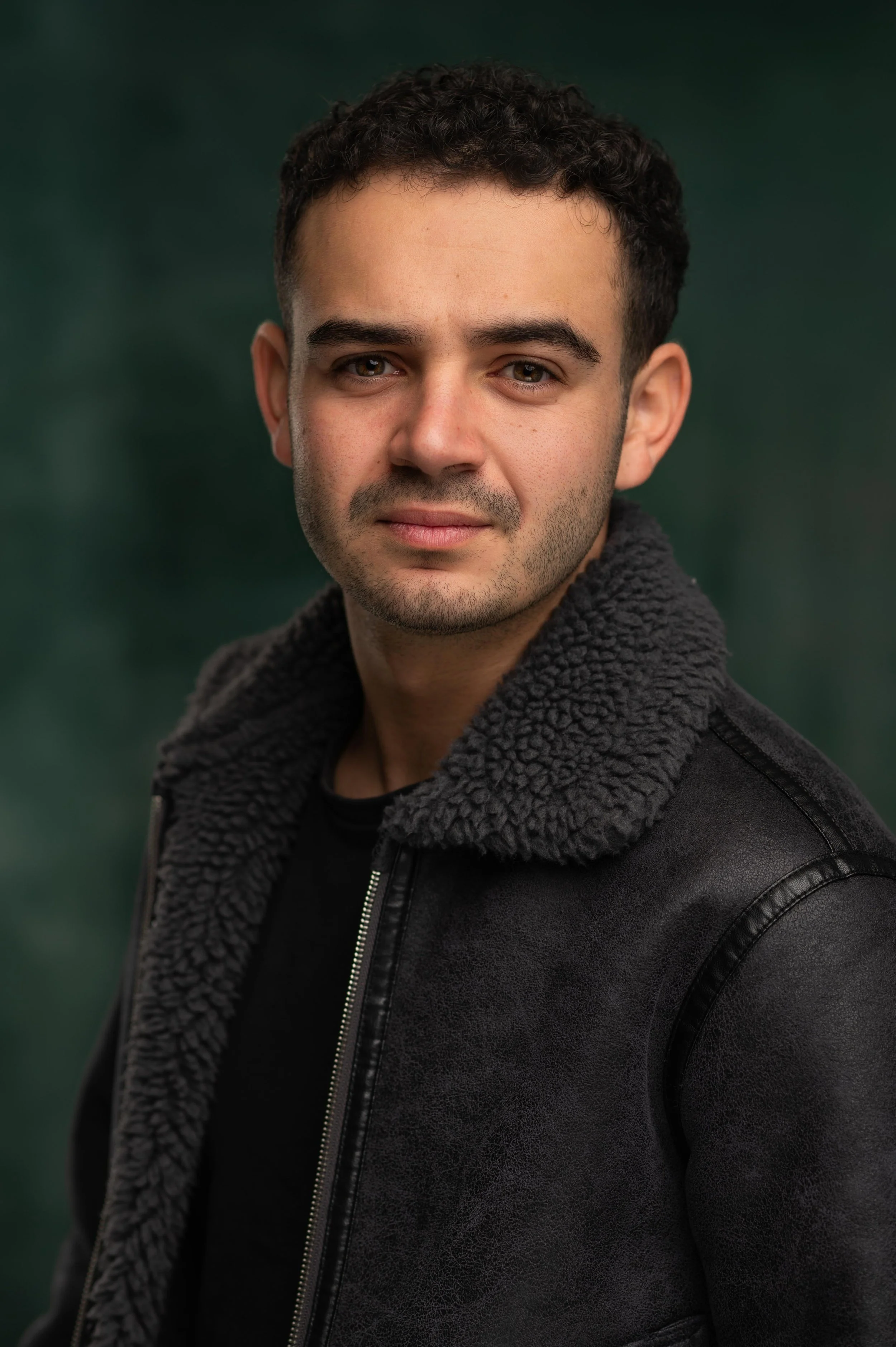 Close-up portrait of a young man with curly dark hair, light skin, and hazel eyes, wearing a black jacket with a gray fleece collar, against a blurred green background.