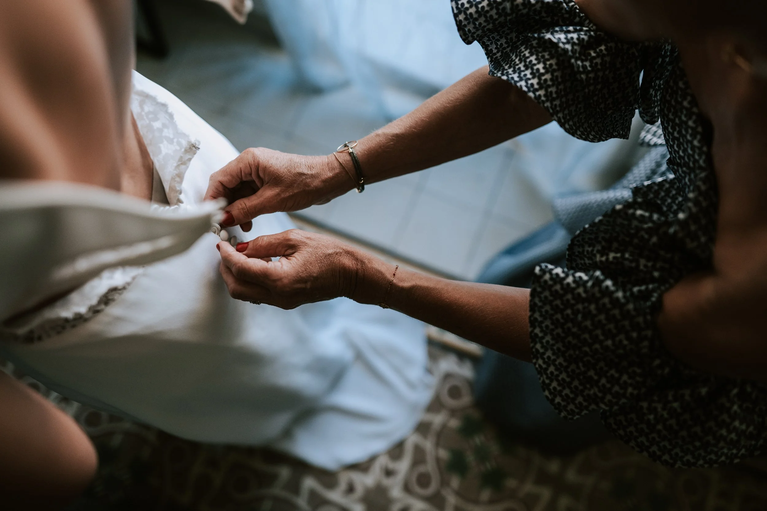 A person helping another person with a white lace gown, possibly a wedding dress, with their hands. The background shows a tiled floor and part of a patterned rug.