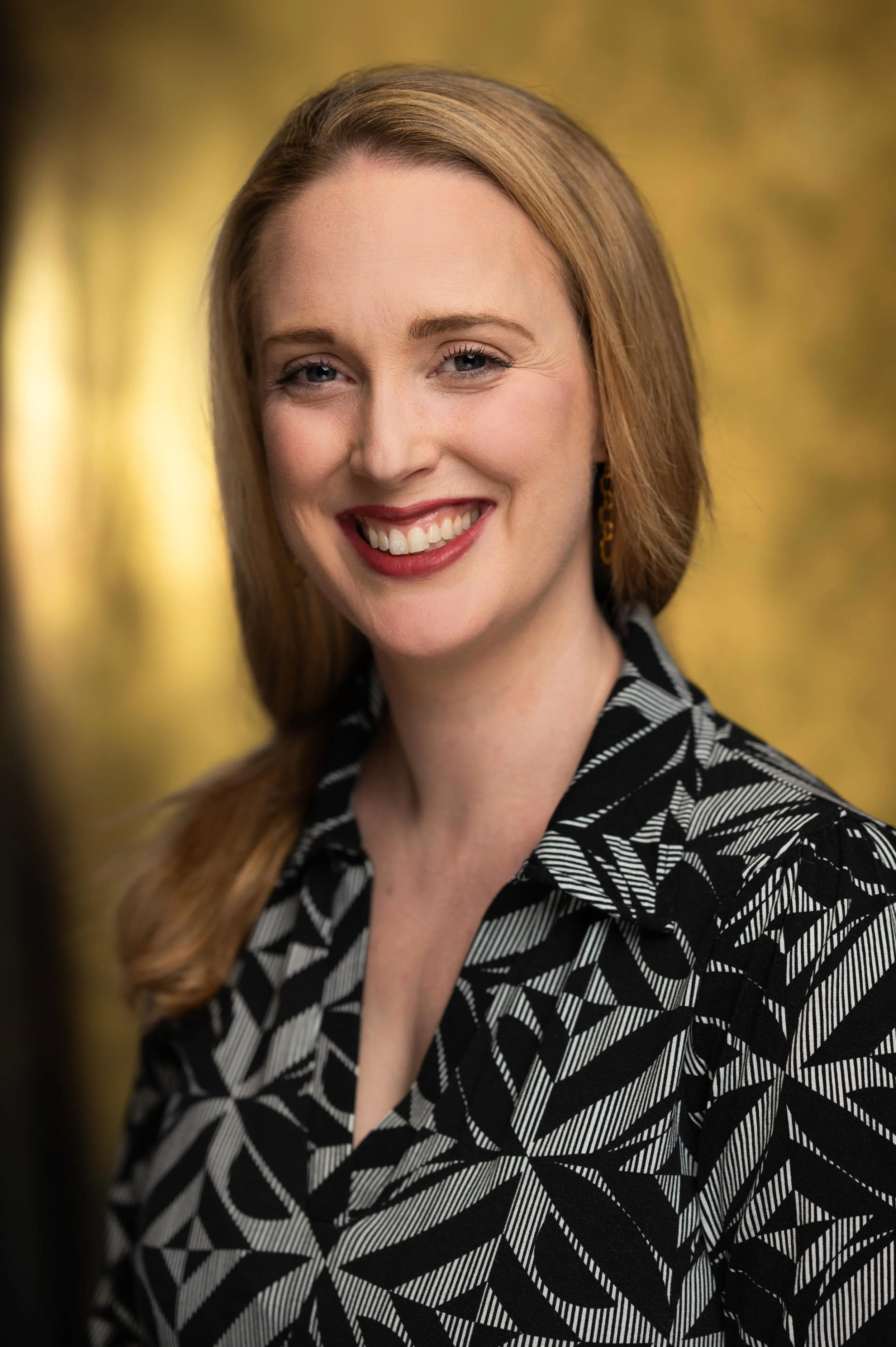 A woman with long red hair, fair skin, and blue eyes, smiling in front of a blurred yellow and green background, wearing a black and white geometric patterned blouse.