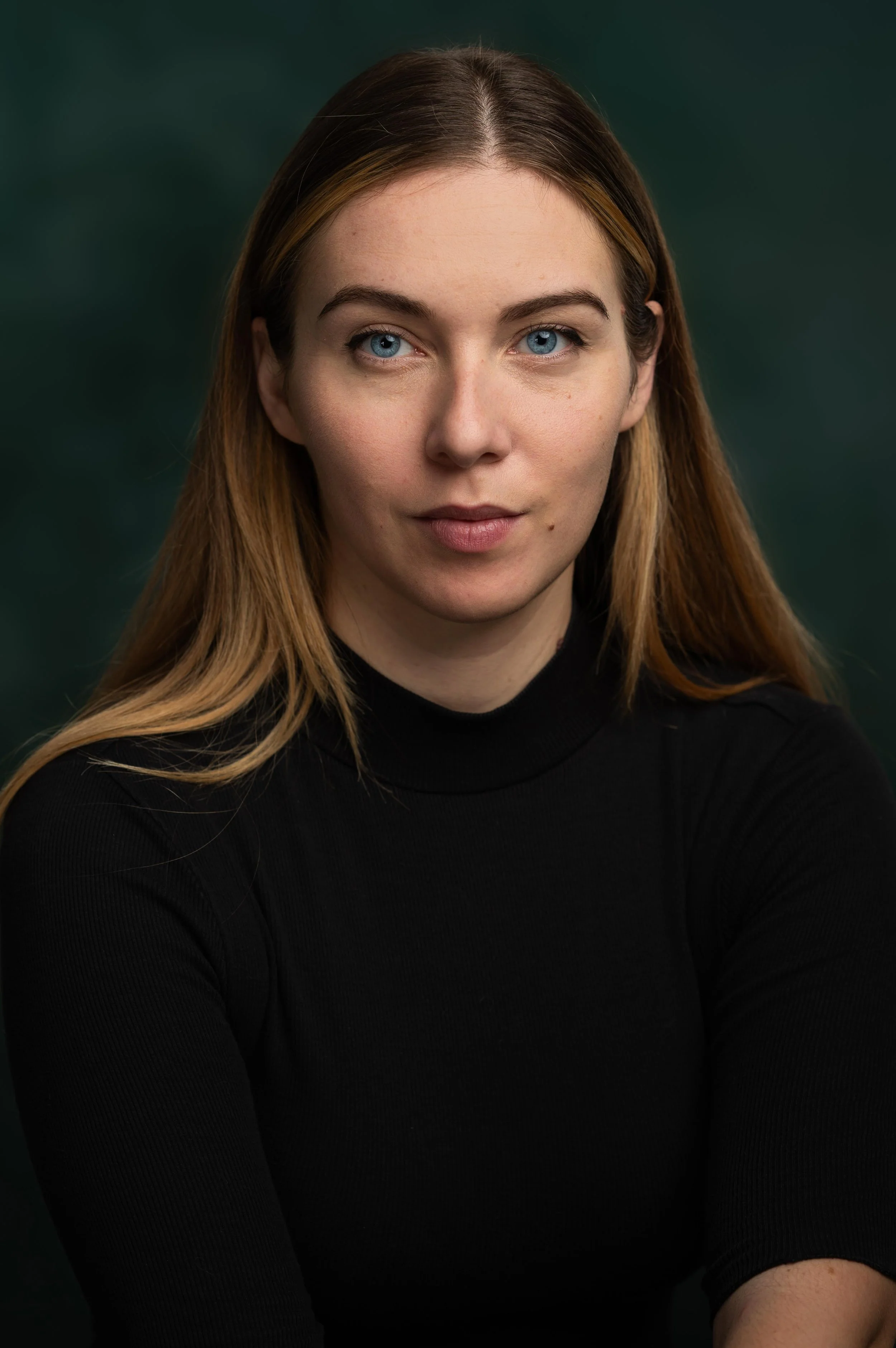 Close-up portrait of a woman with blue eyes and light brown hair, wearing a black turtleneck, against a dark green background.