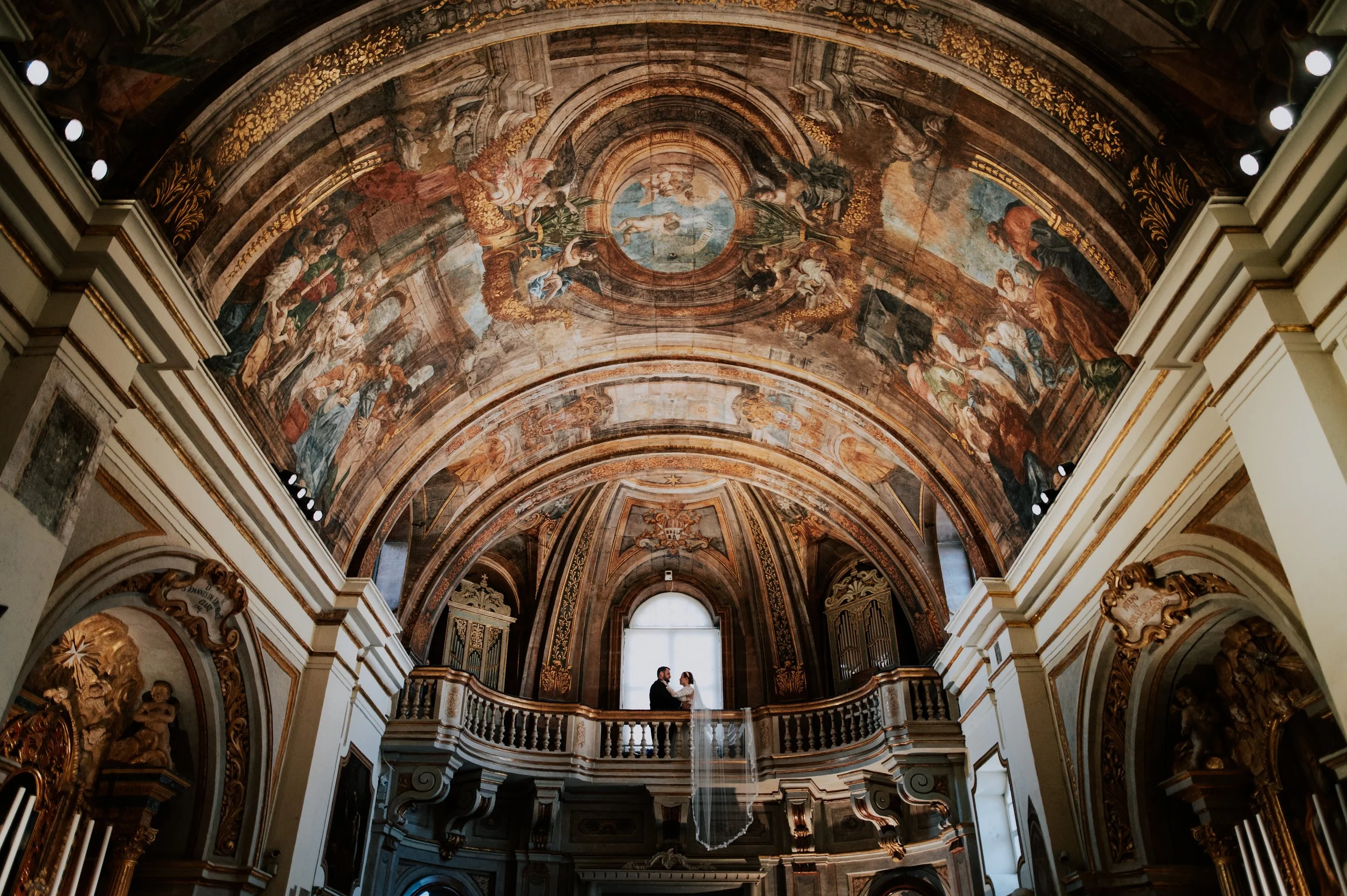 Couple dressed in wedding attire standing on balcony inside a grand, ornately decorated church with a painted vaulted ceiling.