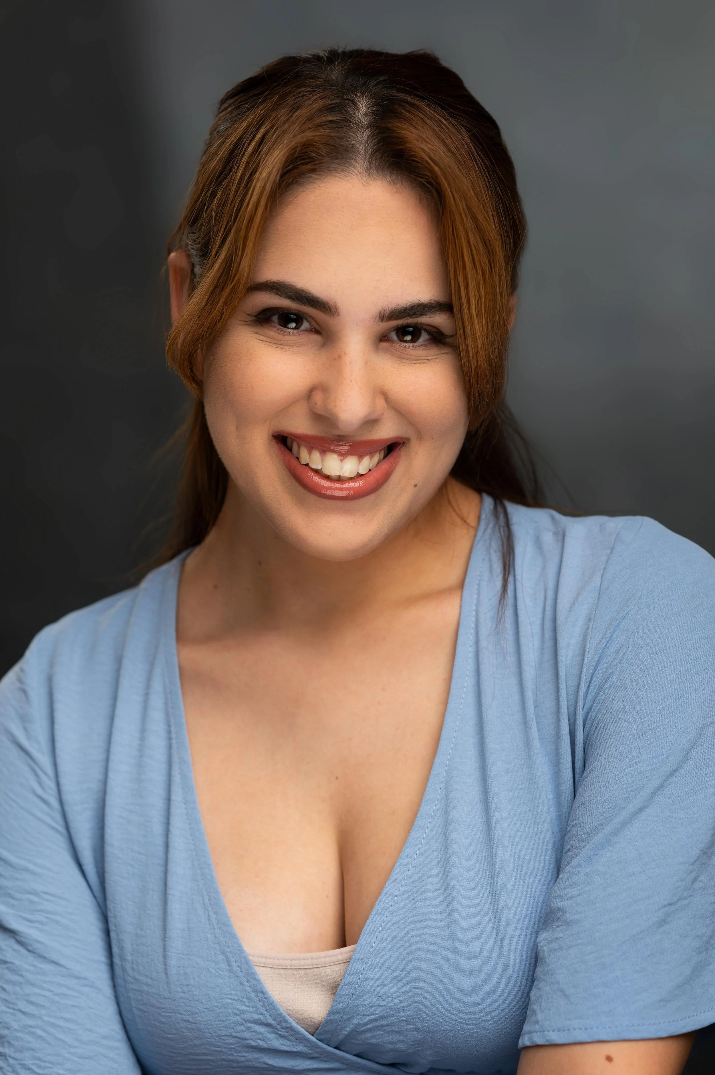 Close-up of a woman smiling with brown hair, wearing a light blue top and beige shirt underneath, against a dark grey background.