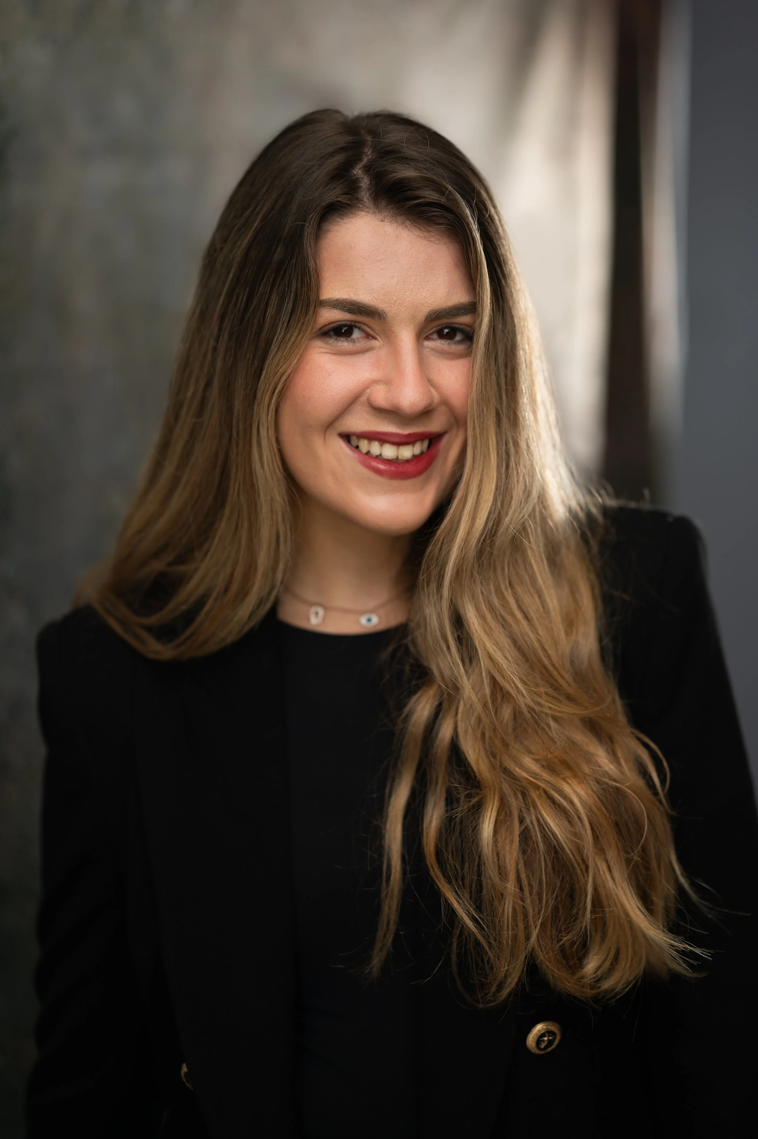 A young woman with long, wavy, light brown hair, smiling, wearing a black blazer with gold buttons and a delicate necklace, standing in front of a blurred background.