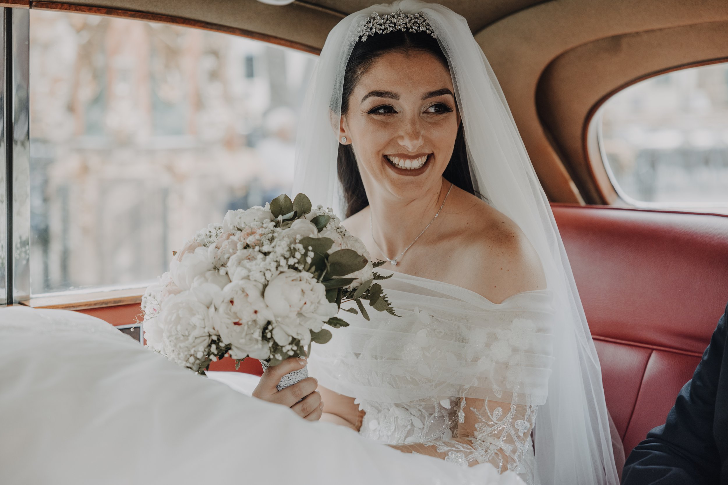 A bride with dark hair and makeup sitting inside a vintage car with wood trim, wearing a white wedding dress and veil, holding a bouquet of white flowers and greenery, smiling and looking to her left.