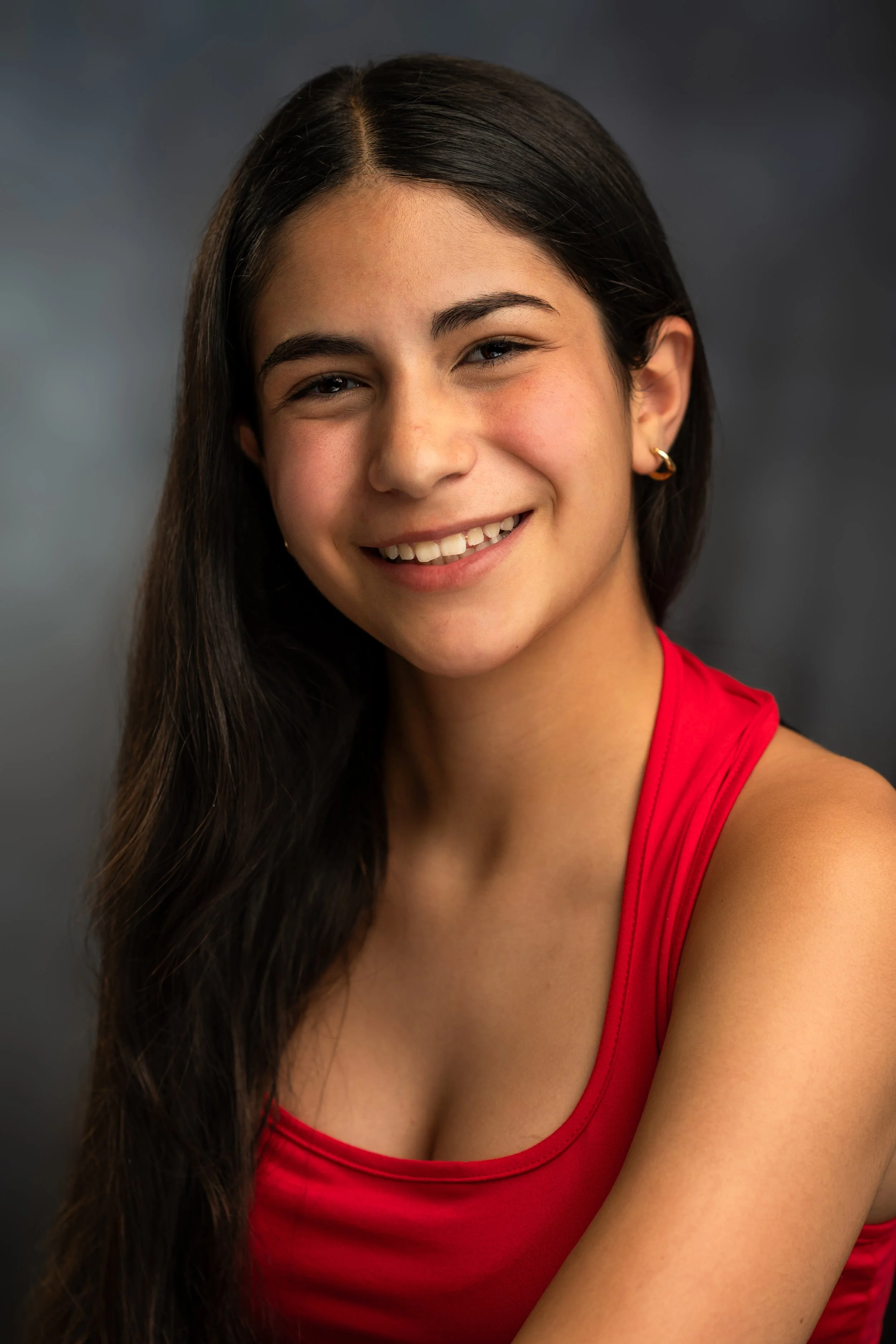 Close-up of a smiling woman with long dark hair, wearing a red sleeveless top and gold hoop earrings, against a dark grey background.
