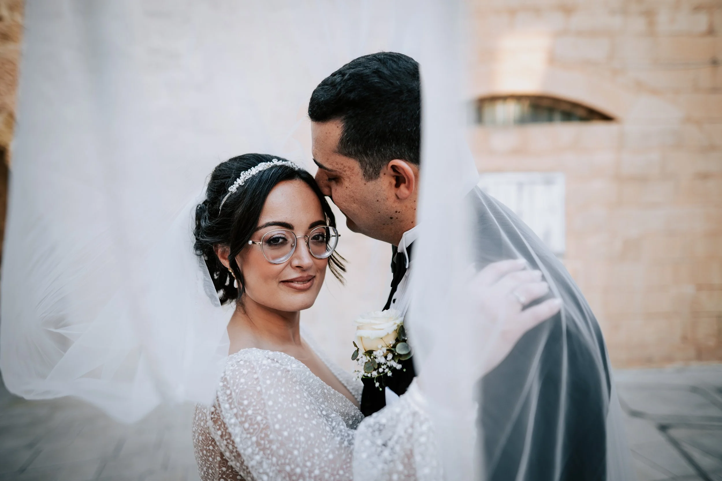 A bride and groom on their wedding day, with the groom gently kissing the bride's forehead as she smiles softly, in a bright indoor setting.