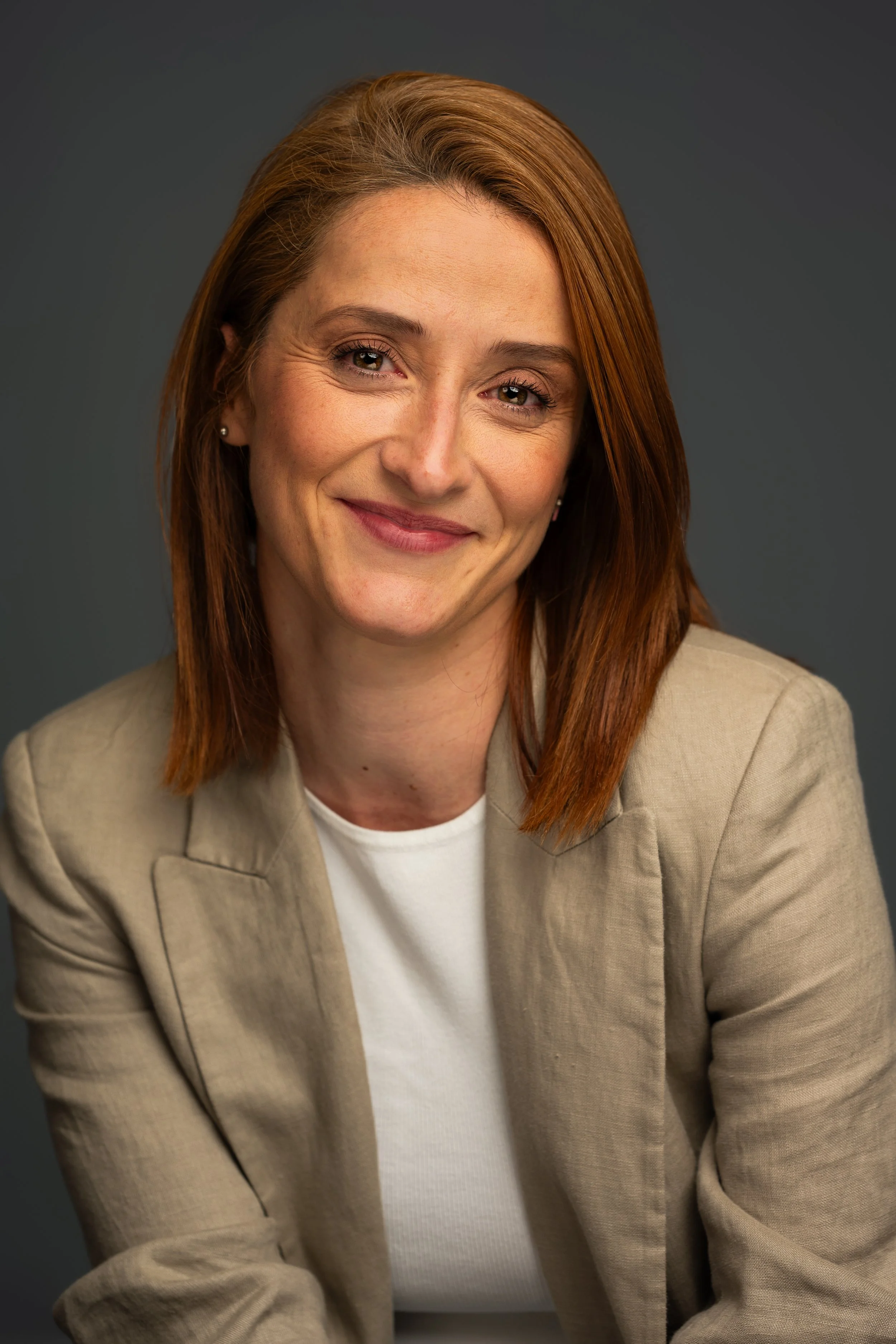A woman with red hair and light skin smiling, wearing a beige blazer over a white shirt, against a gray background.