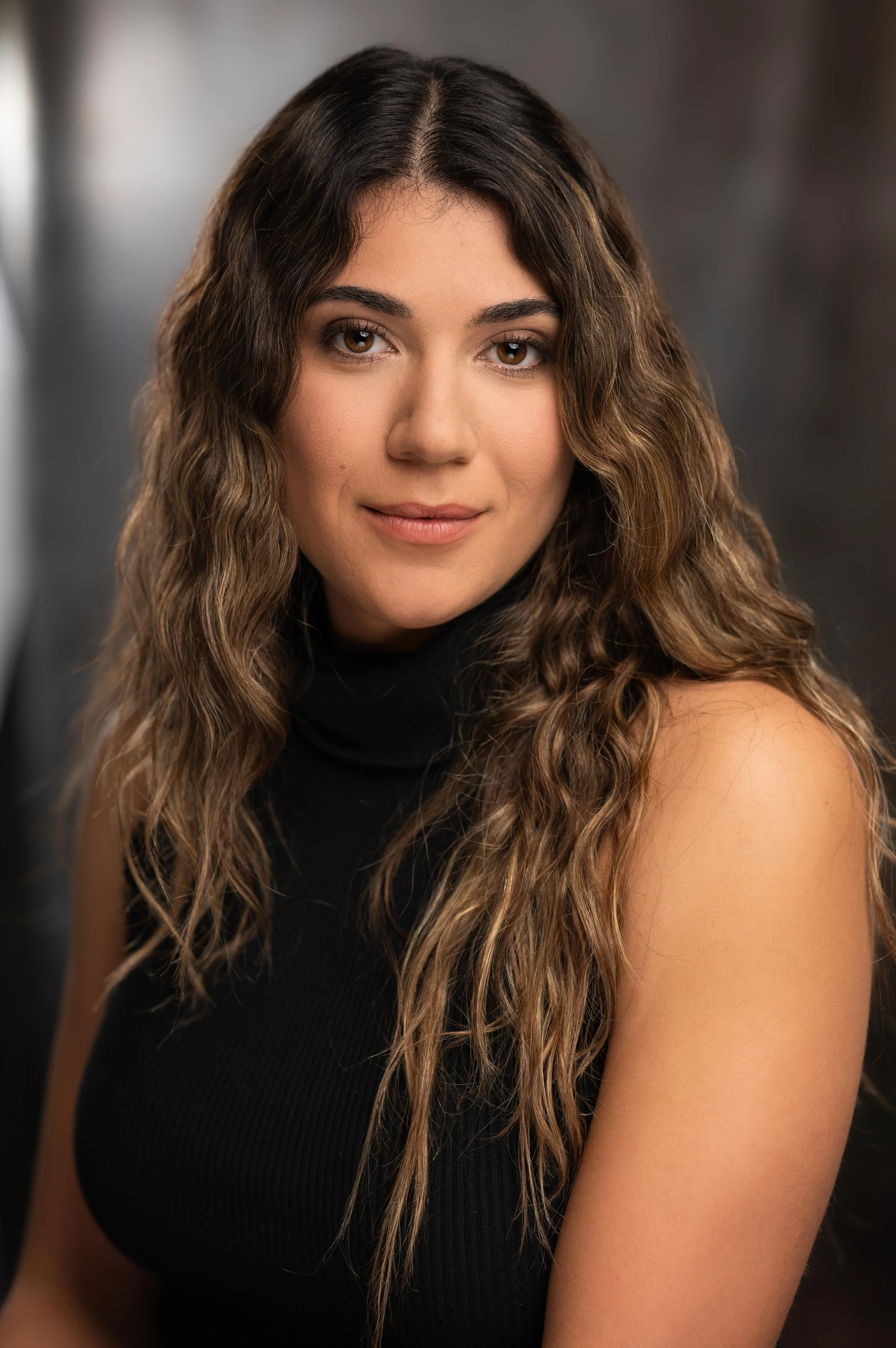 Close-up portrait of a woman with long, wavy brown hair, wearing a black sleeveless top, looking at the camera with a slight smile.