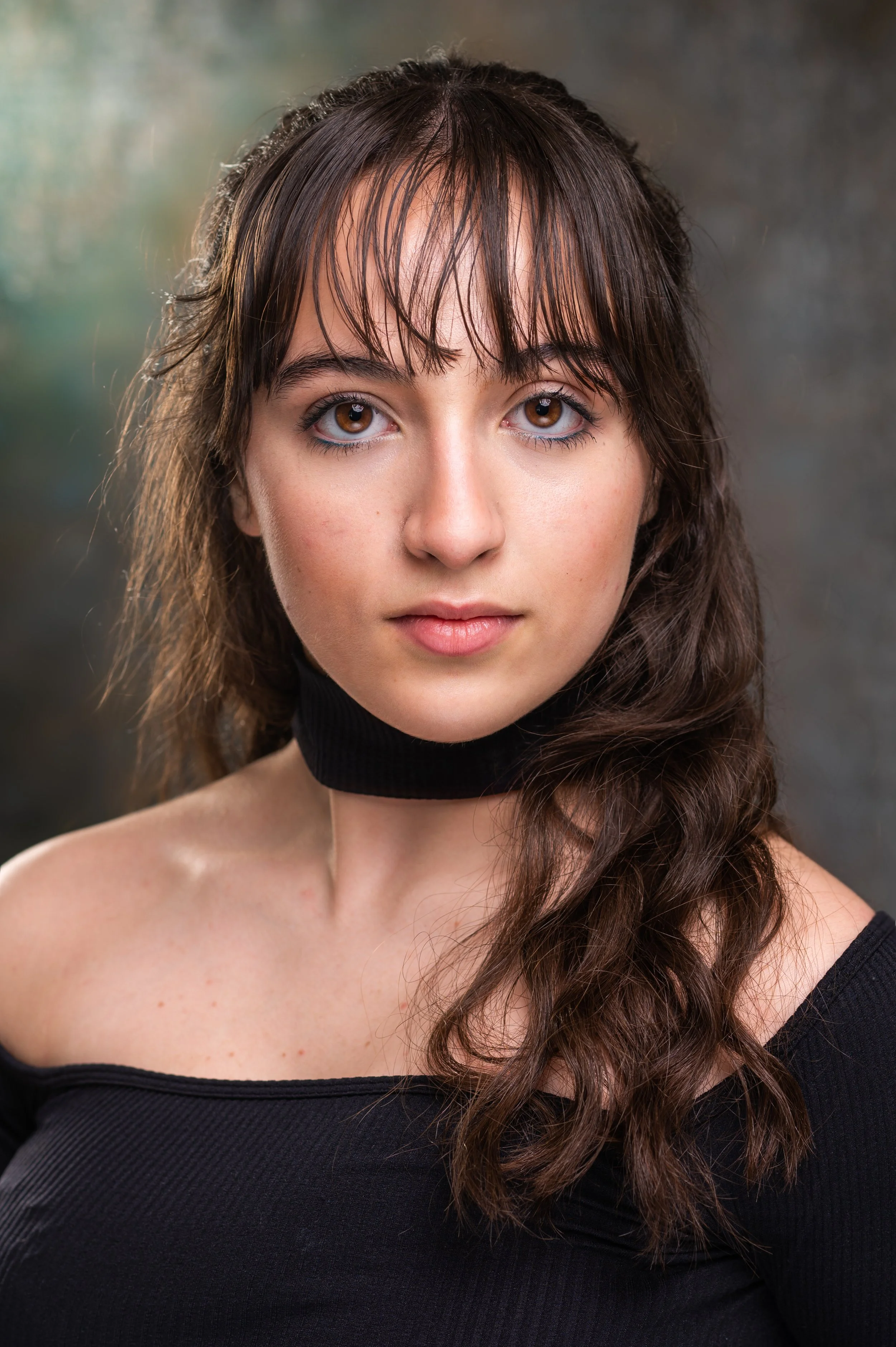 Close-up portrait of a young woman with brown hair, brown eyes, and bangs, wearing a black off-shoulder top and black choker, with a neutral expression, against a blurred green and gray background.