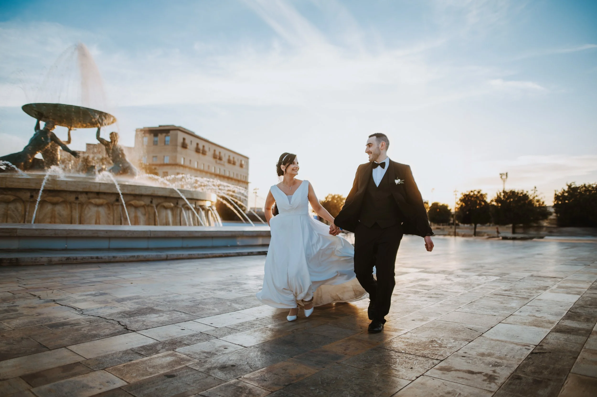 A bride and groom holding hands and walking in front of a fountain at sunset.