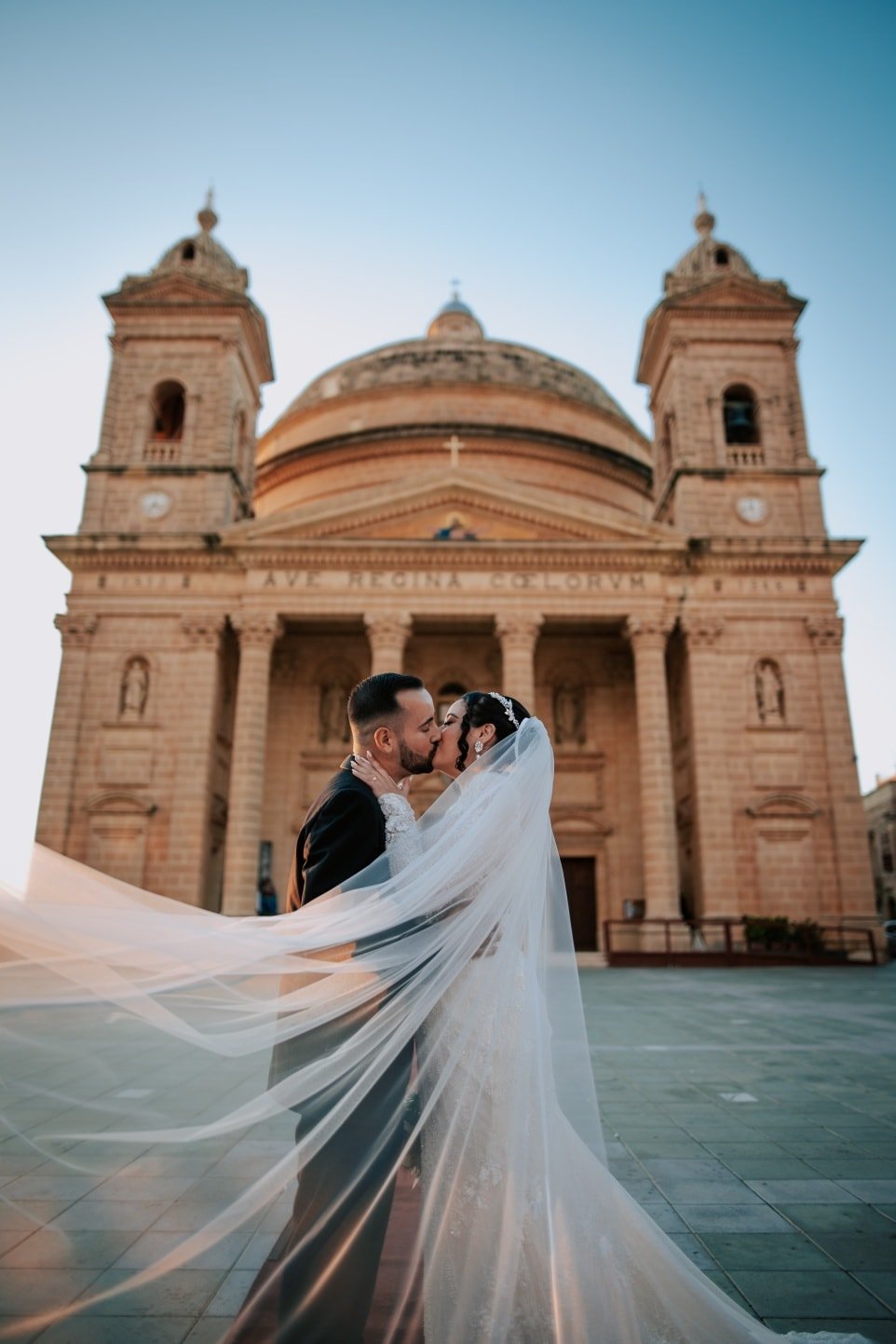 A bride and groom sharing a kiss in front of a historic church, with the bride’s veil flowing in the wind during sunset.