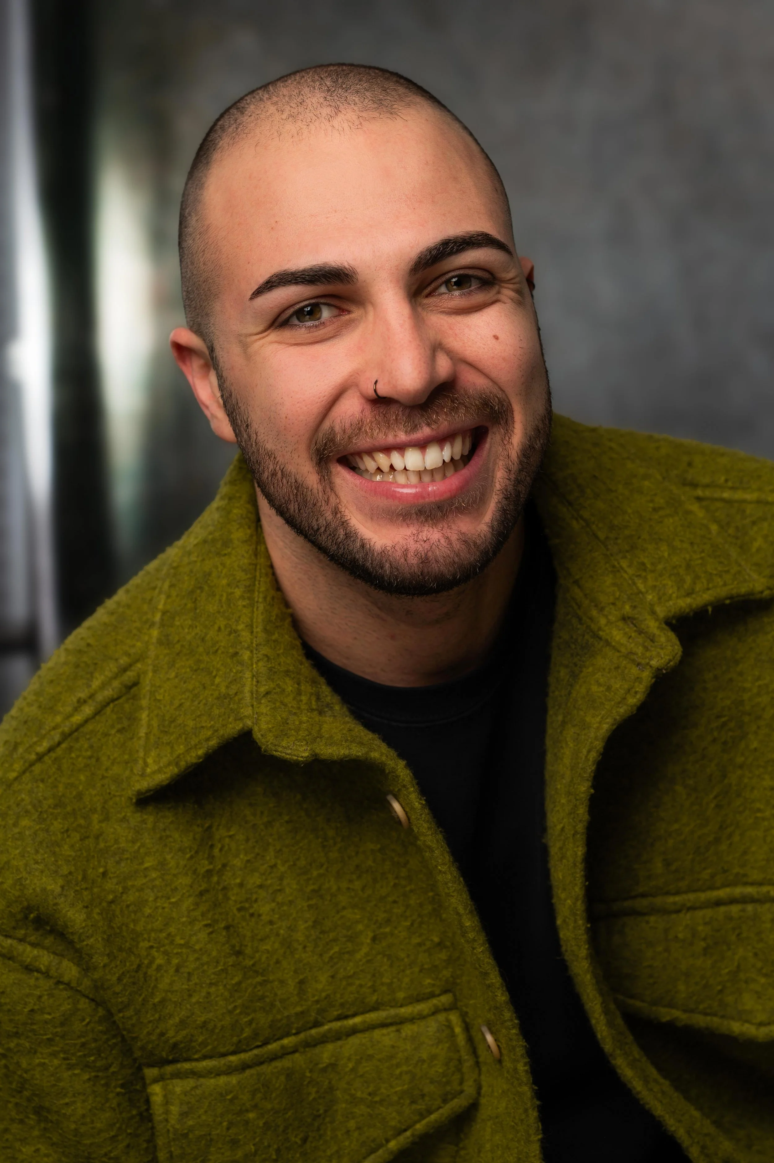 Smiling young man with a beard, nose ring, and short hair wearing a green jacket.