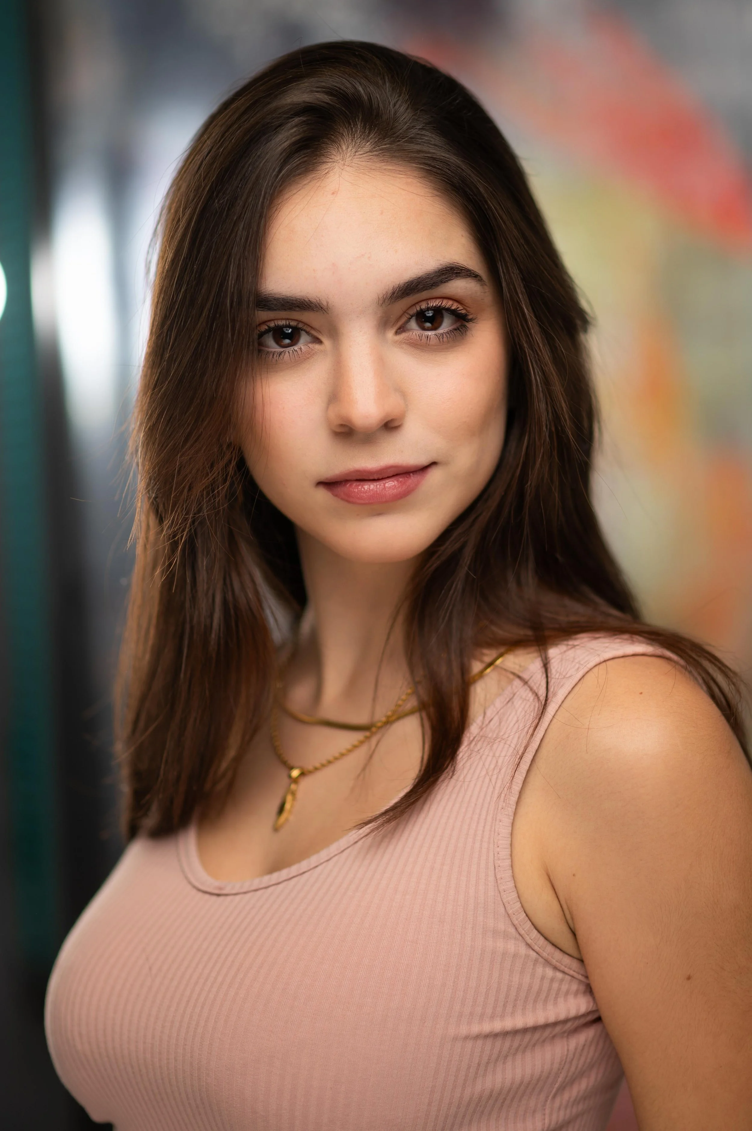Close-up of a young woman with long brown hair, brown eyes, wearing a pink tank top and layered gold necklaces, standing against a blurred background.