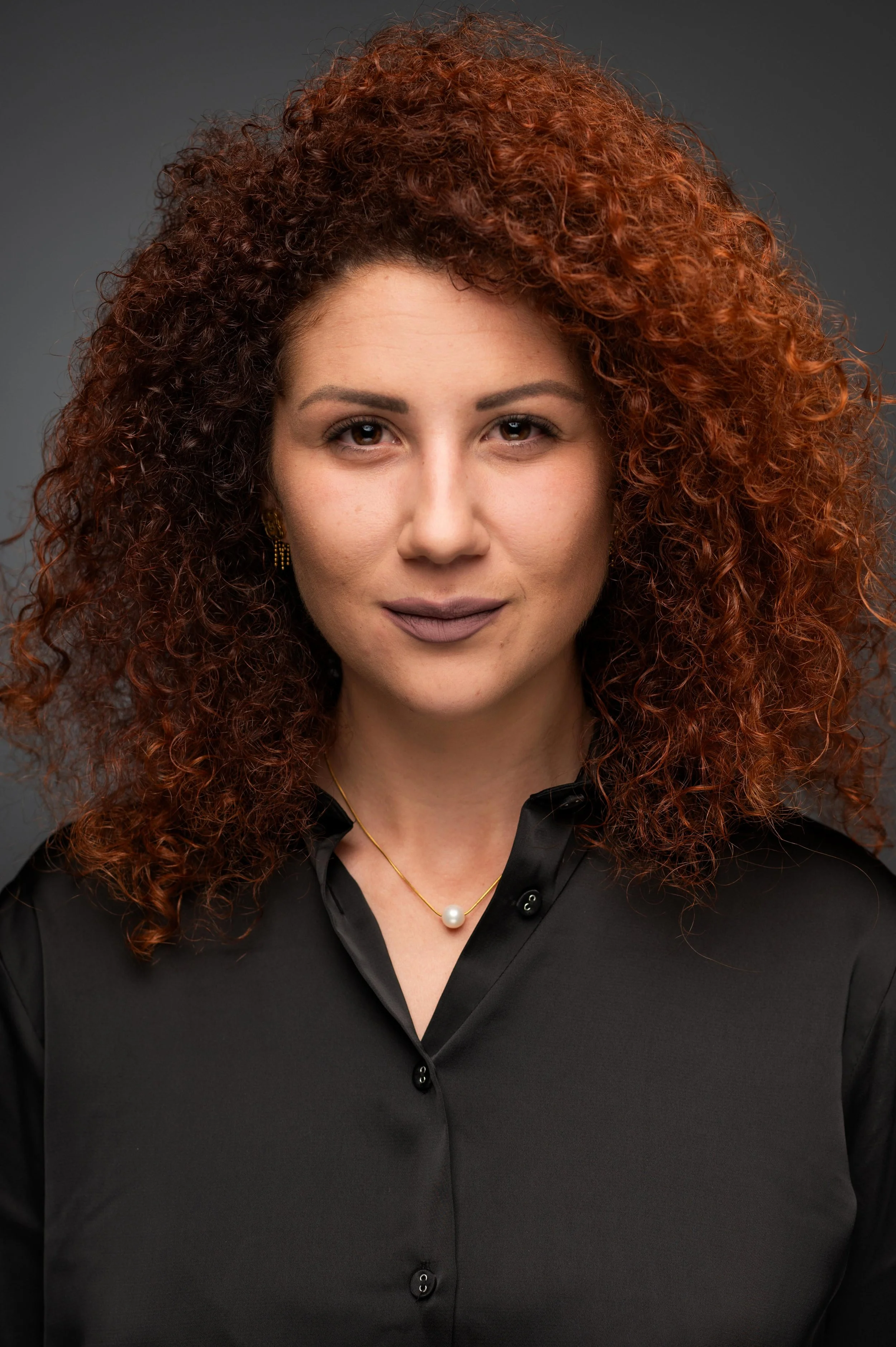 A woman with curly red hair wearing a black blouse, gold earrings, and a necklace with a pearl pendant poses against a dark gray background.