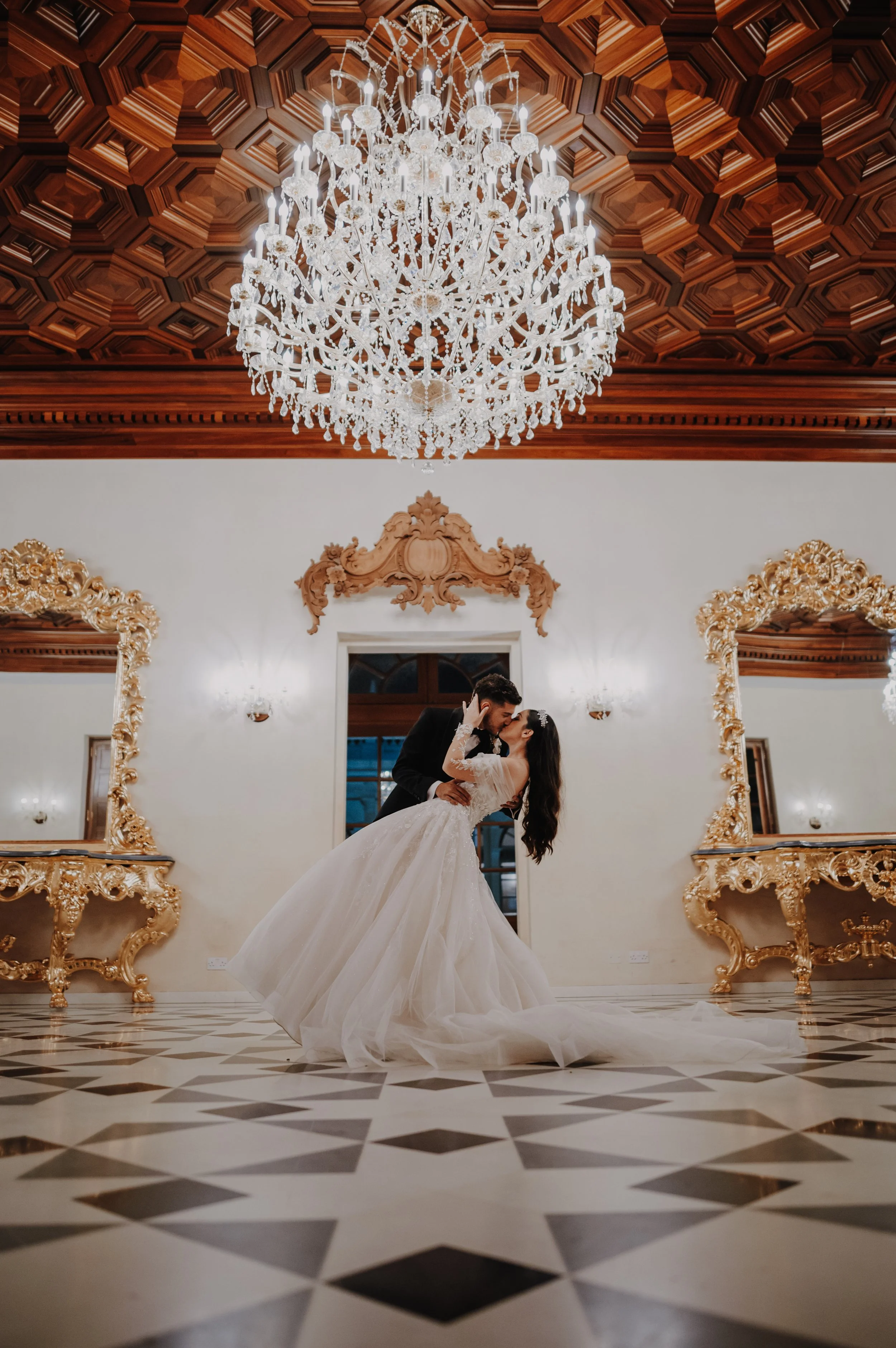 A bride and groom sharing a romantic dance in a luxurious, ornate room with a large crystal chandelier overhead and gold-trimmed mirrors adorning the walls. The bride is in a flowing white wedding gown, and the groom is in a black suit.