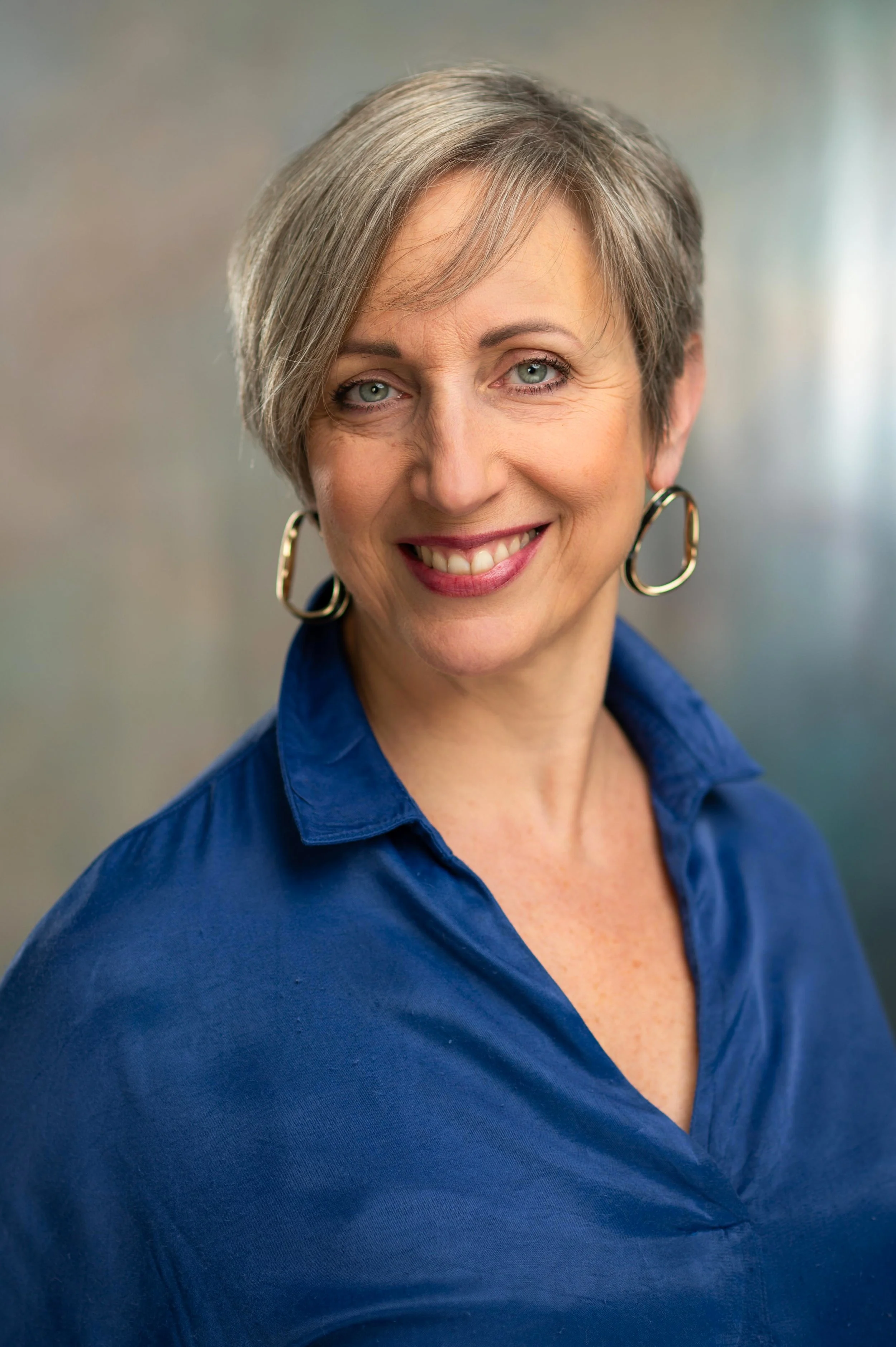 A woman with short, gray hair, blue eyes, wearing hoop earrings and a blue shirt, smiling at the camera.