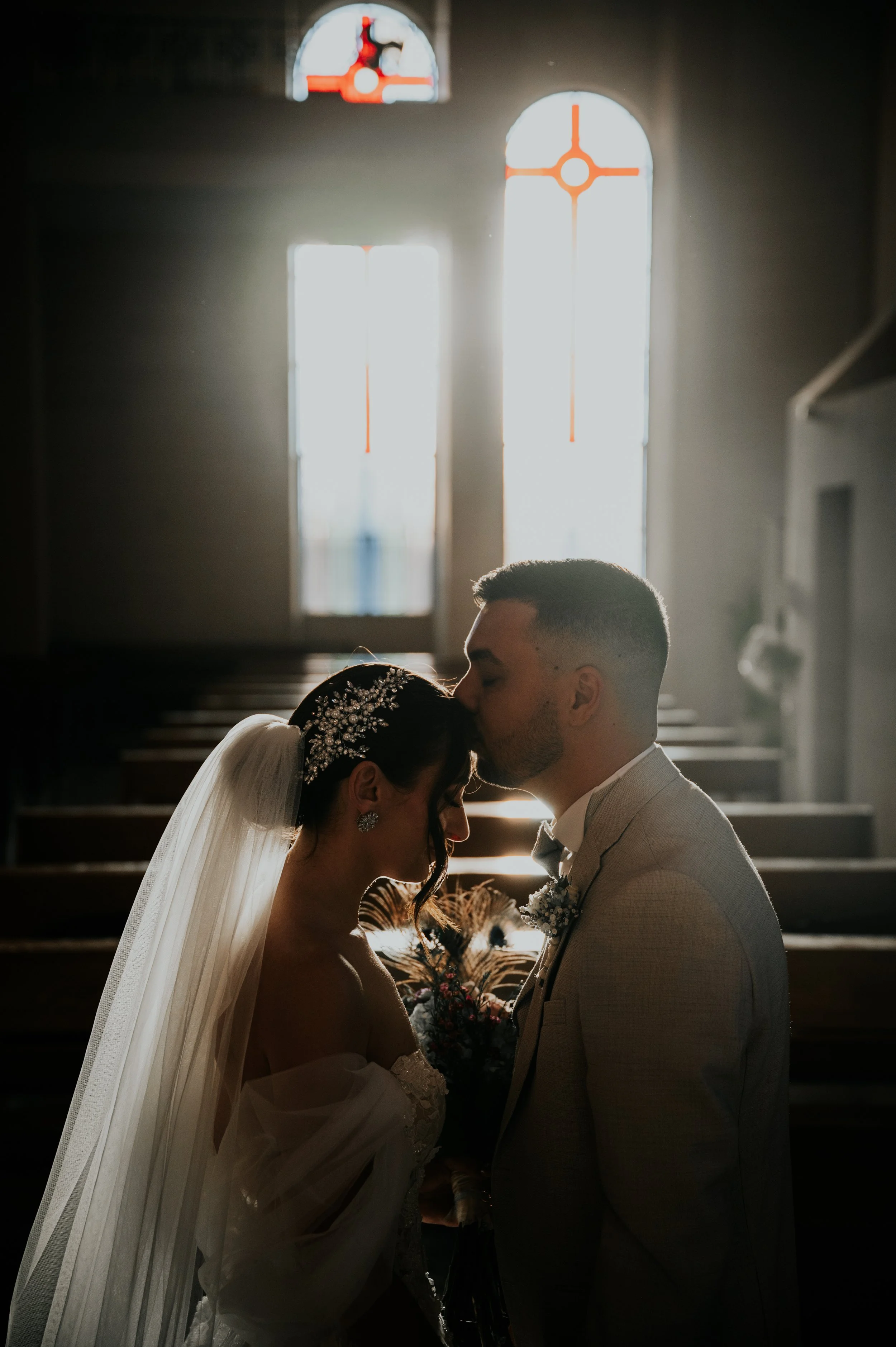 A bride and groom sharing a kiss inside a church with large arched windows backlit by sunlight.