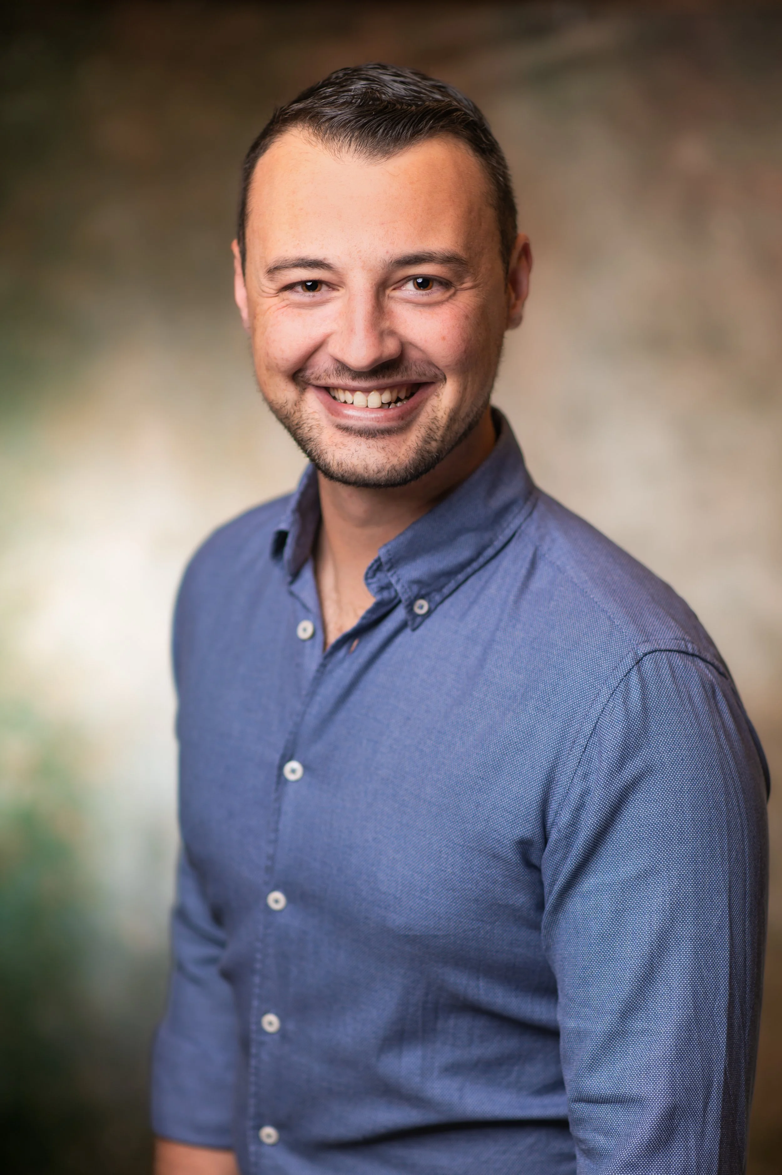 Headshot of Matthew Muscat Drago smiling, wearing a blue button-up shirt, against a blurred outdoor background.