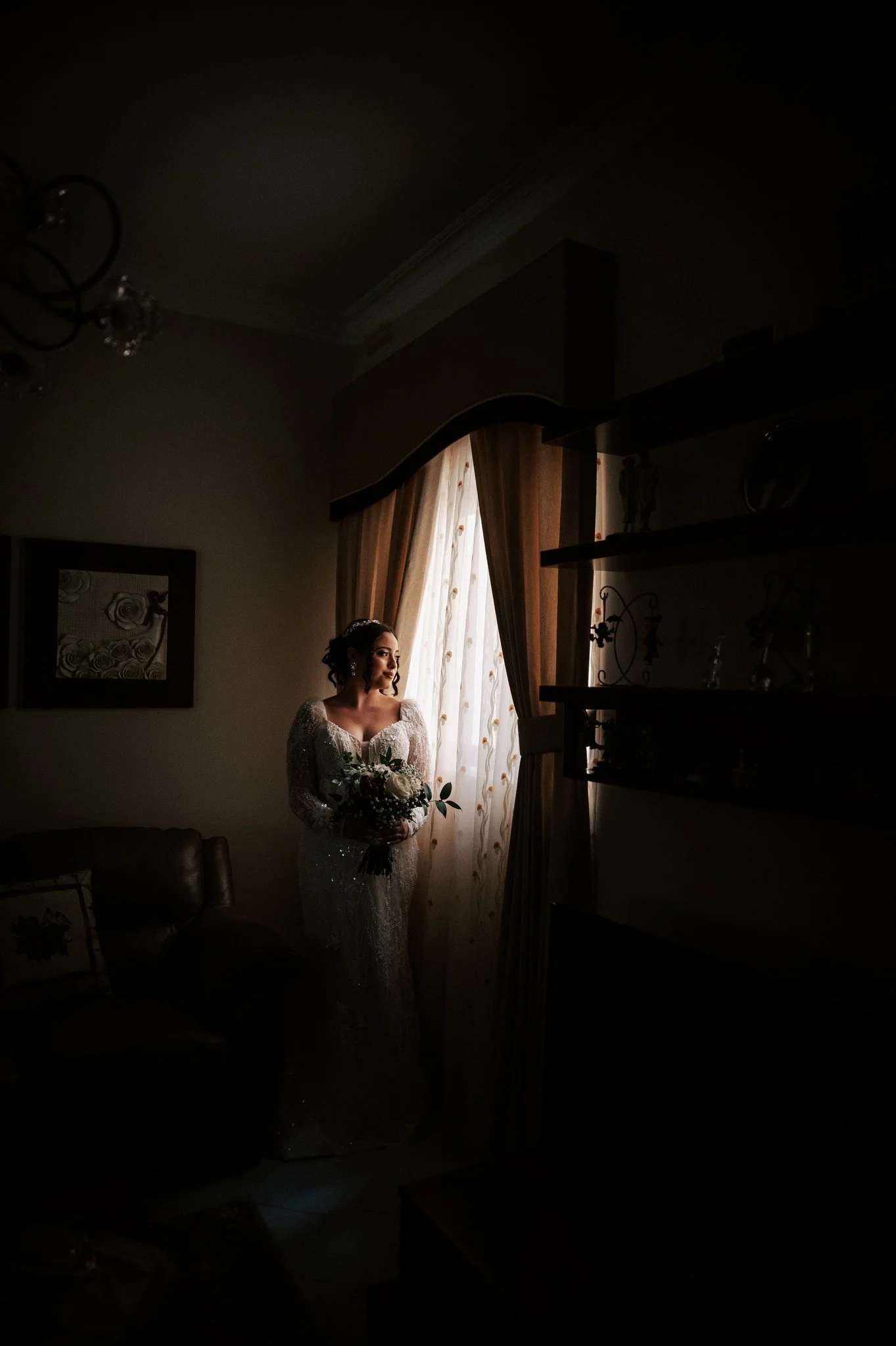 A woman in a white dress holding a bouquet, standing by a window with light curtains, in a dimly lit room.