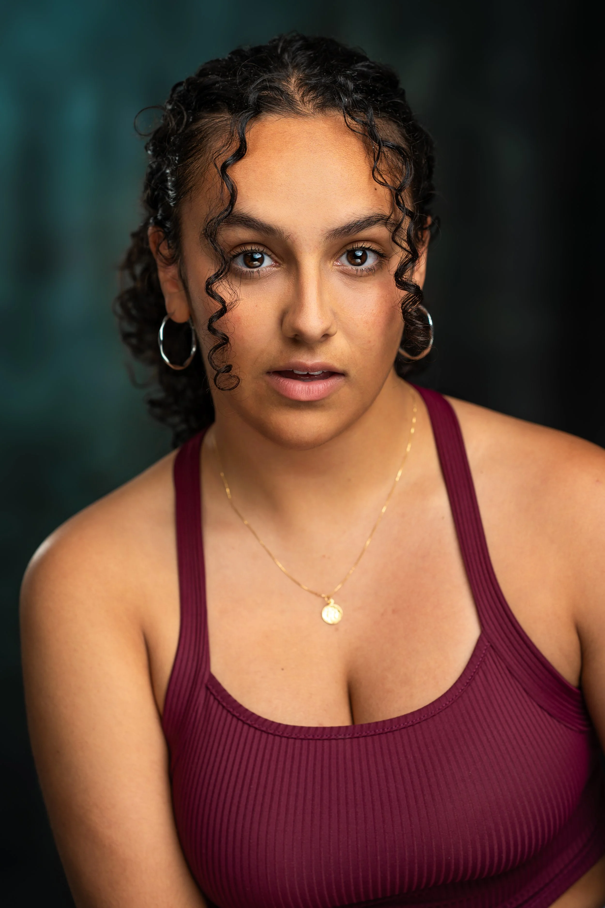 Close-up portrait of a young woman with dark curly hair, wearing hoop earrings and a burgundy tank top, looking directly into the camera with a neutral expression against a dark background.
