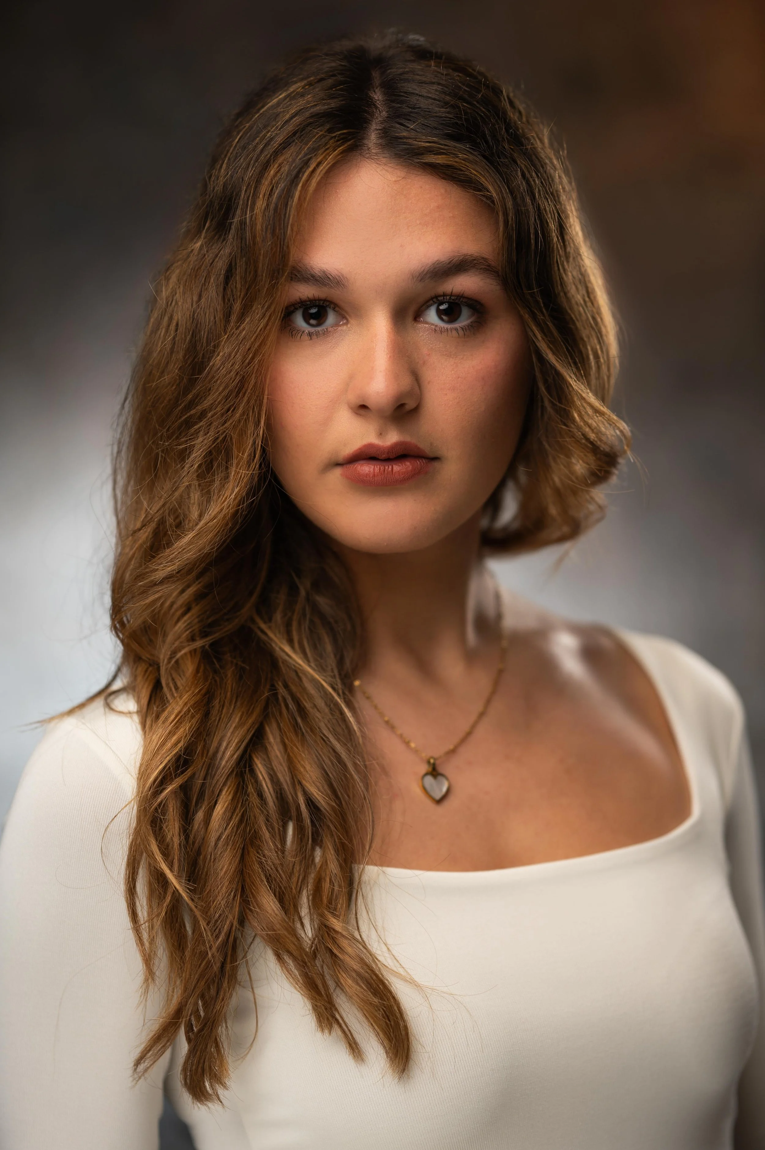 A young woman with long, wavy brown hair, wearing a white top, gold necklace with a heart-shaped pendant, looking directly at the camera with a neutral expression.