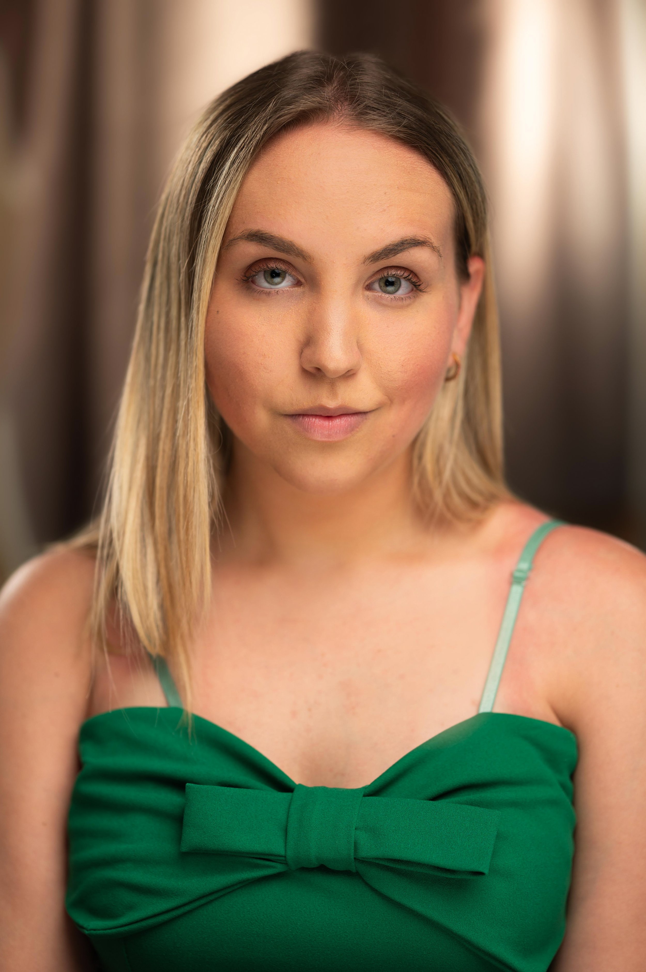 A young woman with long blonde hair and blue eyes wearing a green dress with spaghetti straps and bow detail, standing indoors in front of blurred curtains.