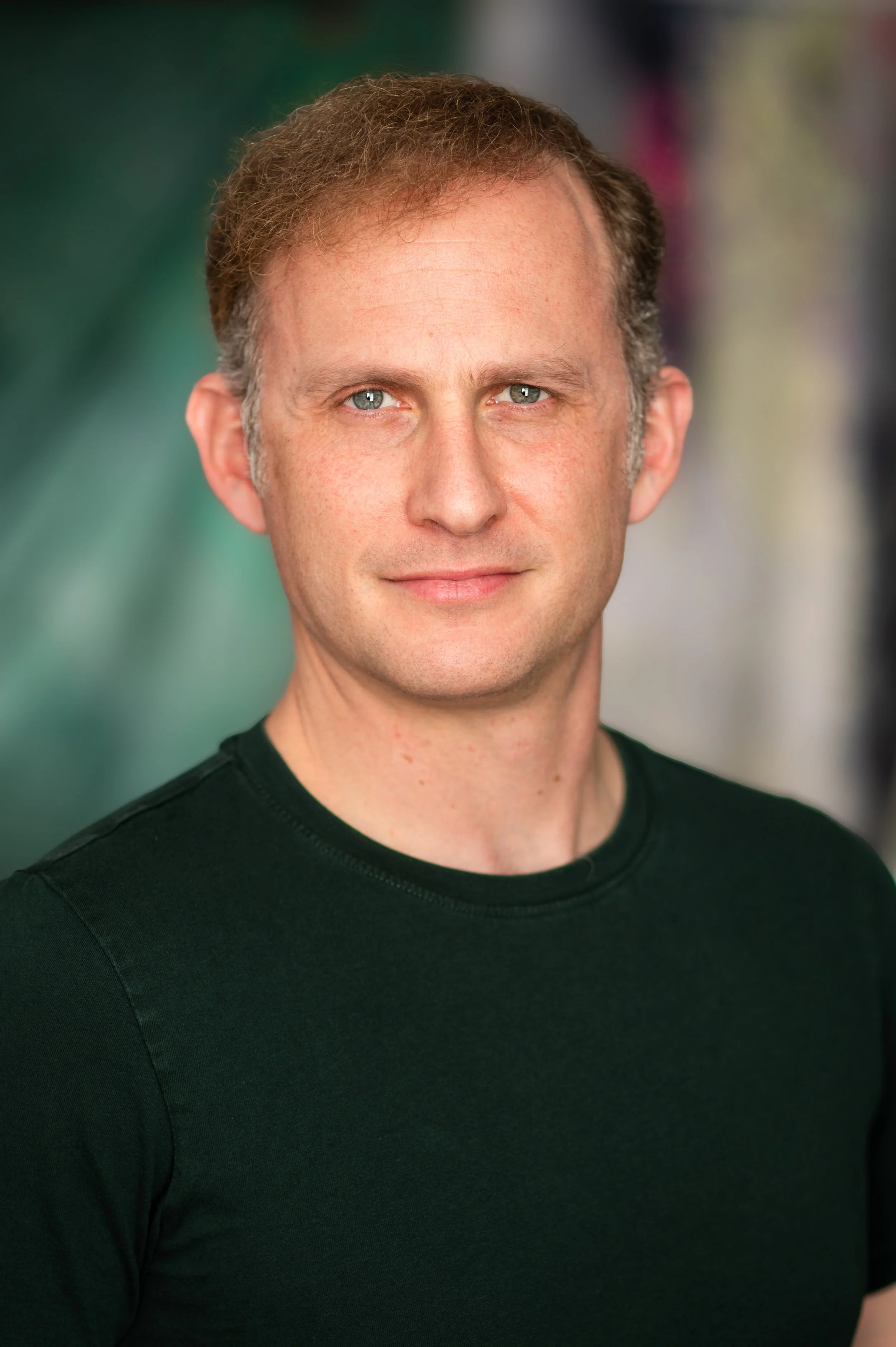 Close-up portrait of a middle-aged man with short, light brown hair and blue eyes, wearing a black t-shirt, against a blurred background.