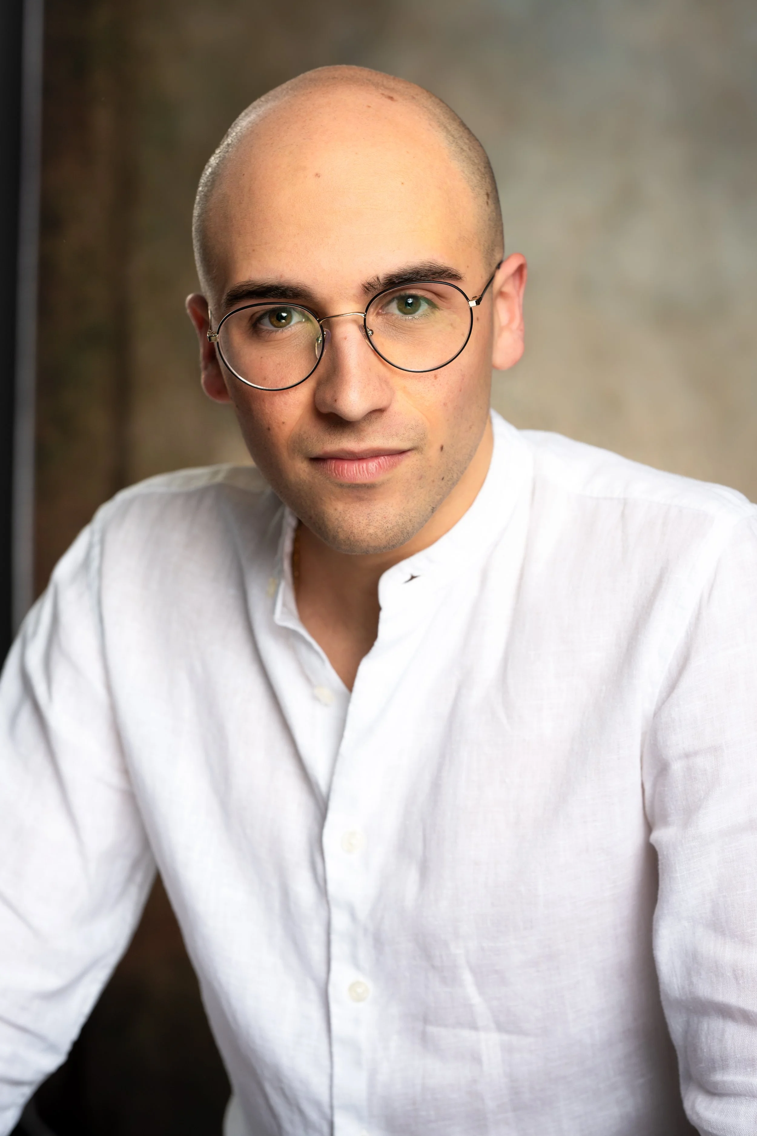 A young man with a bald head, wearing round glasses and a white dress shirt, sitting in front of a neutral background.