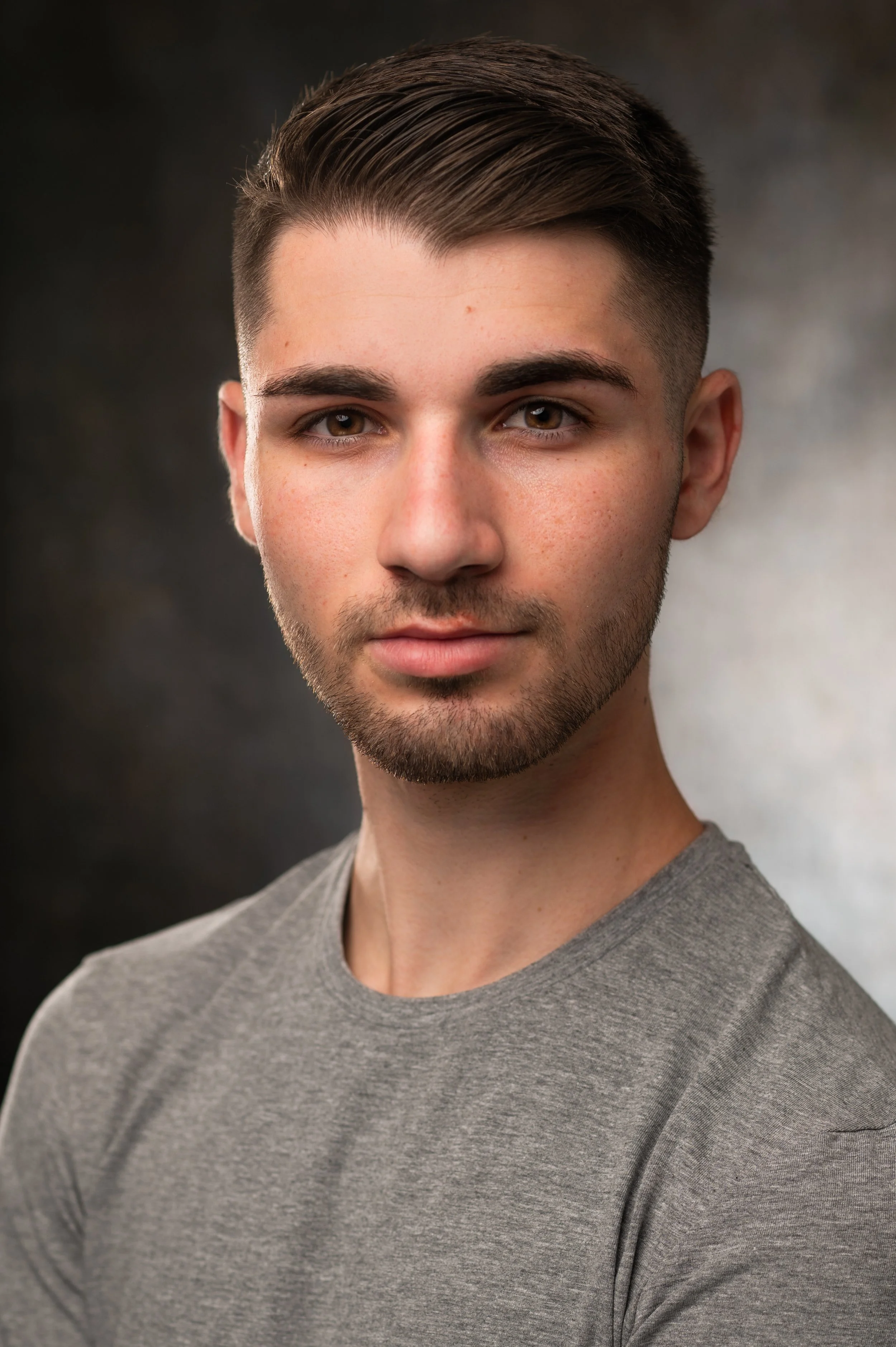 Close-up of a young man with short brown hair, brown eyes, light skin, and a slight beard, wearing a gray T-shirt, against a blurred dark background.