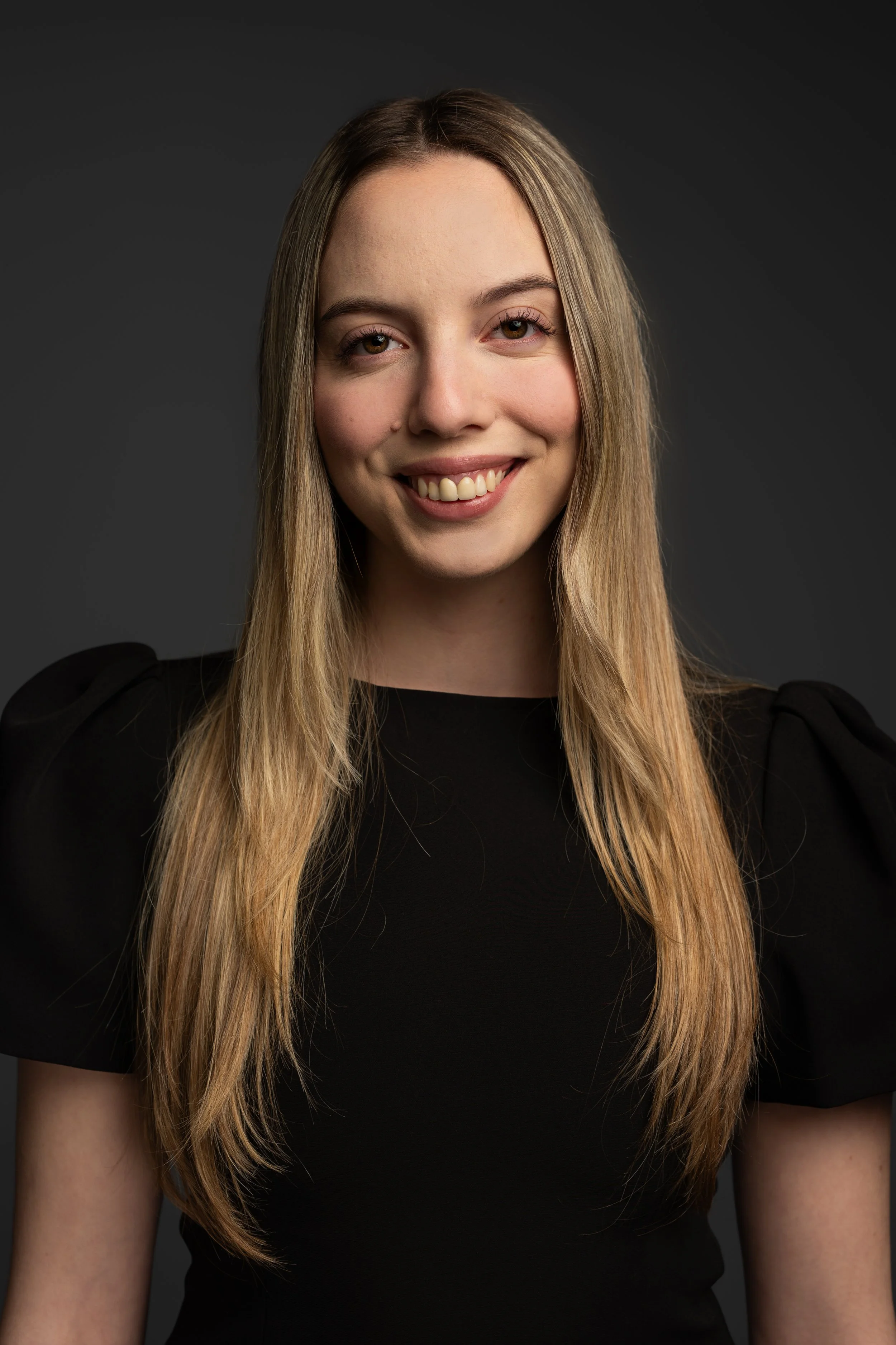 Close-up of a young woman with long blonde hair, smiling, wearing a black top, against a dark background.