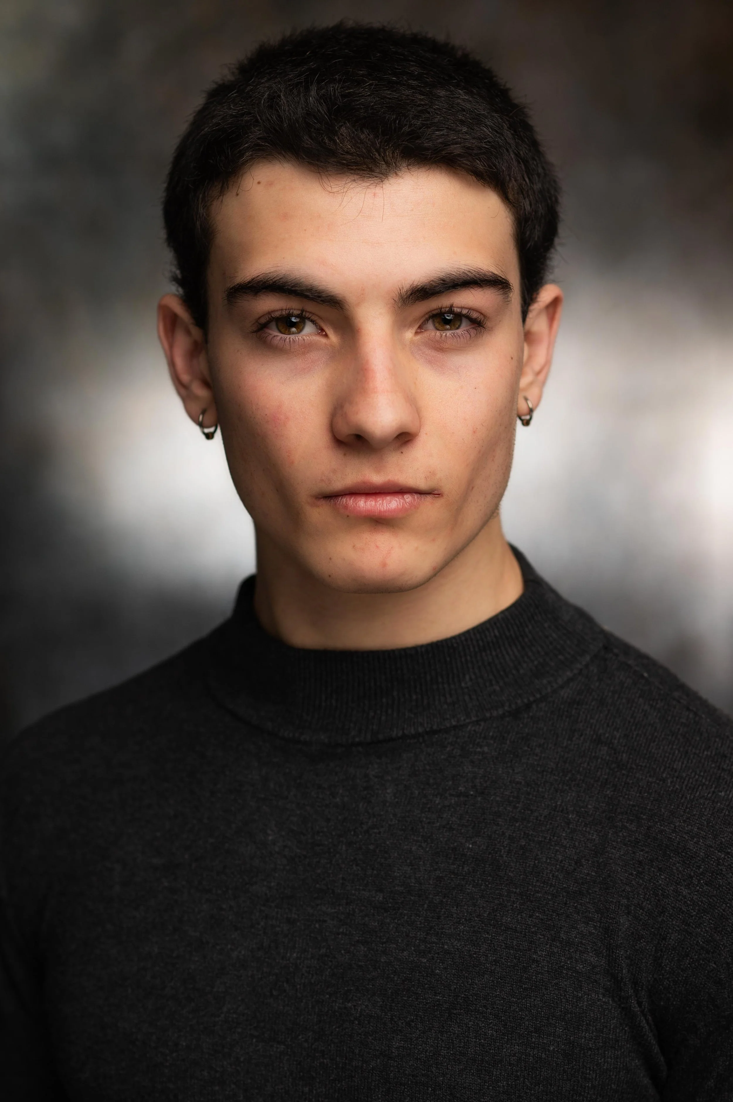A young person with short dark hair, blue eyes, and earrings, wearing a black turtleneck sweater against a blurred dark background.