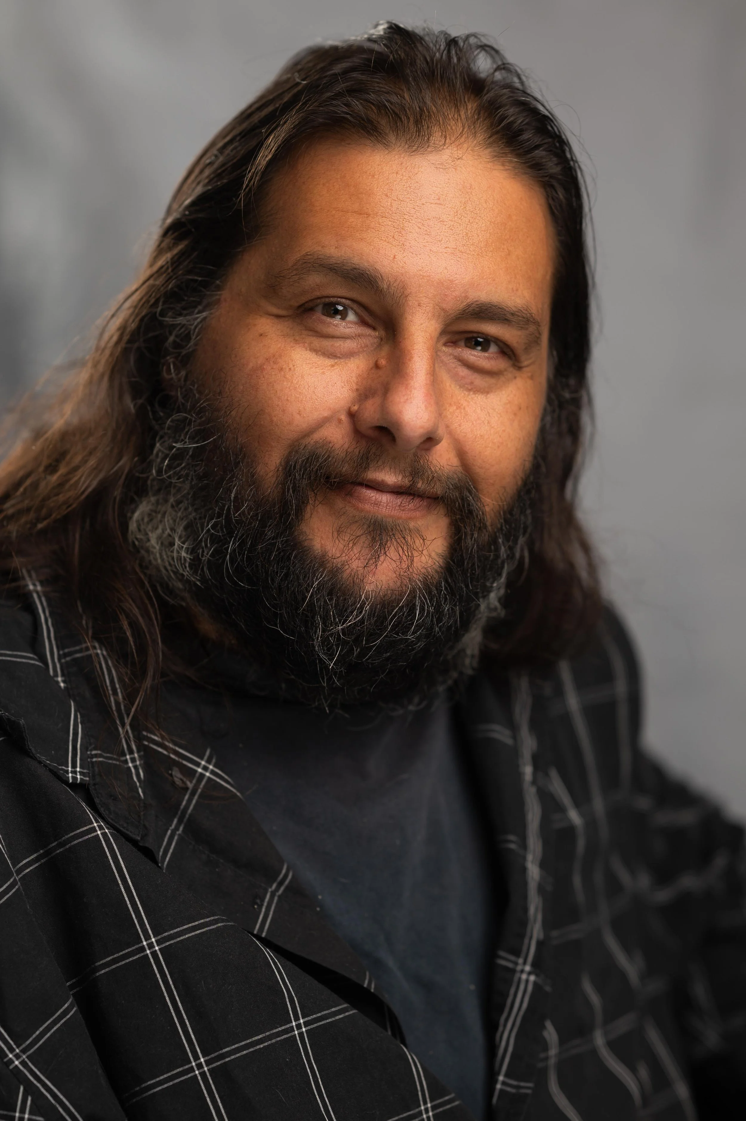 A middle-aged man with long dark hair, a full beard, and a mustache is smiling softly at the camera. He is wearing a black shirt with white pinstripes and a dark t-shirt underneath. The background is a plain, neutral gray.