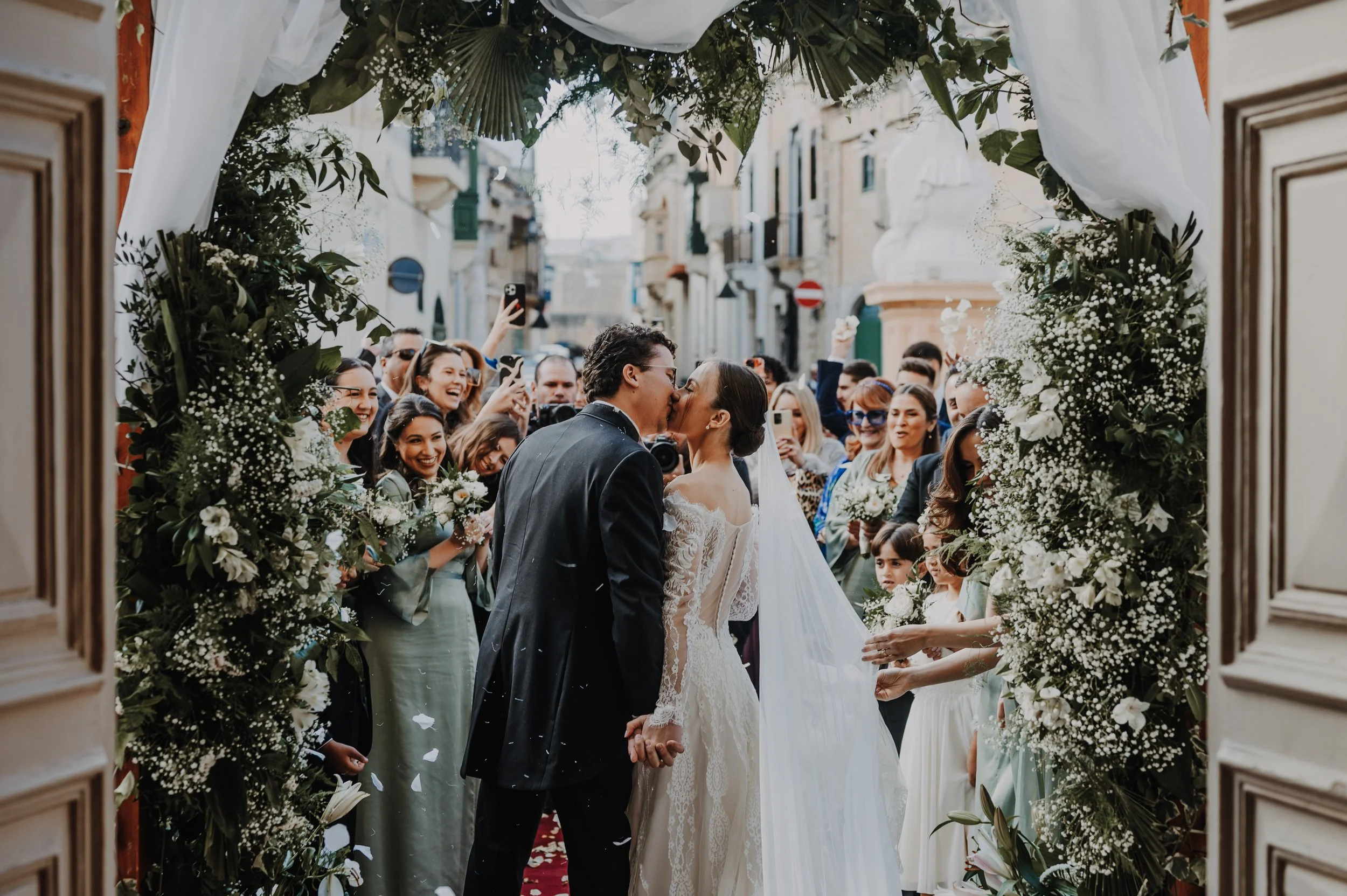A couple sharing a kiss during their wedding ceremony, surrounded by guests, with floral decorations framing the scene.