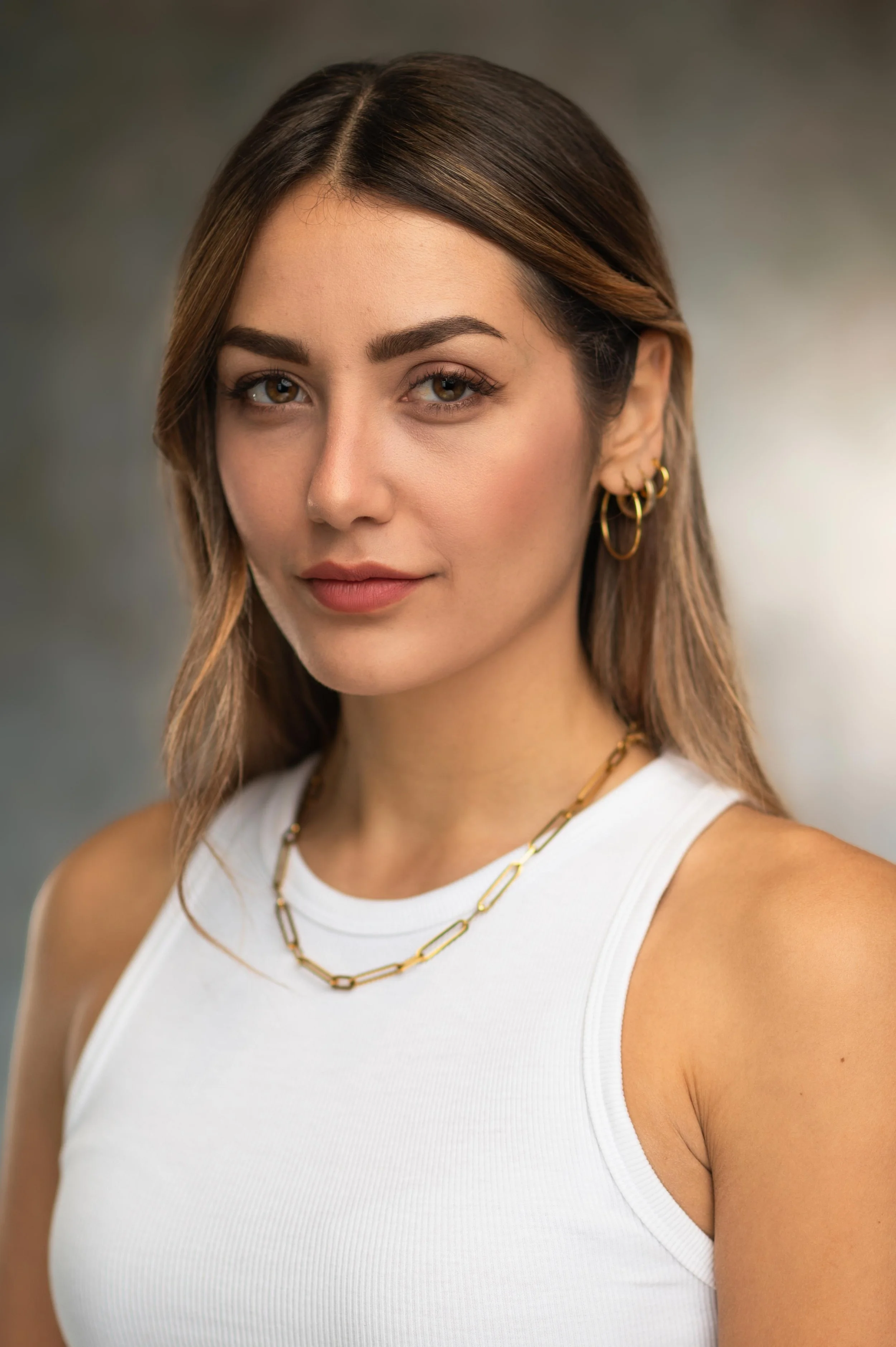 Close-up portrait of a young woman with light brown hair, wearing a white sleeveless top, gold hoop earrings, and gold necklace, smiling softly against a blurred background.