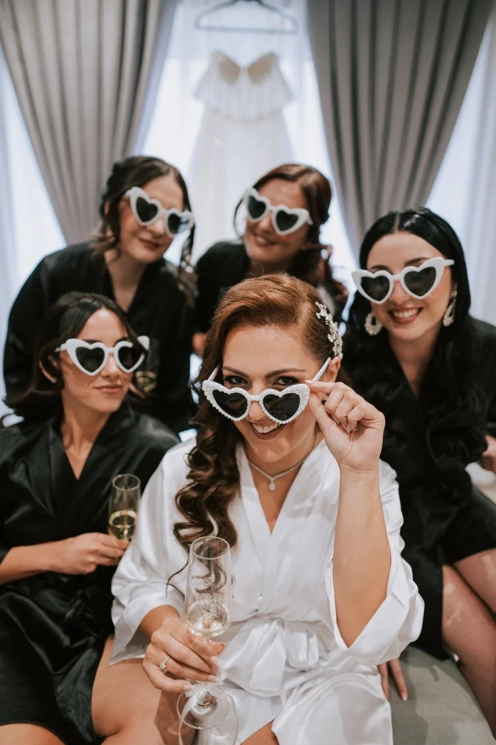 A group of five women, including a bride in a white robe, posing and smiling while wearing heart-shaped sunglasses at a celebration in a wedding suite. The bride is holding a glass of champagne and adjusting her sunglasses.