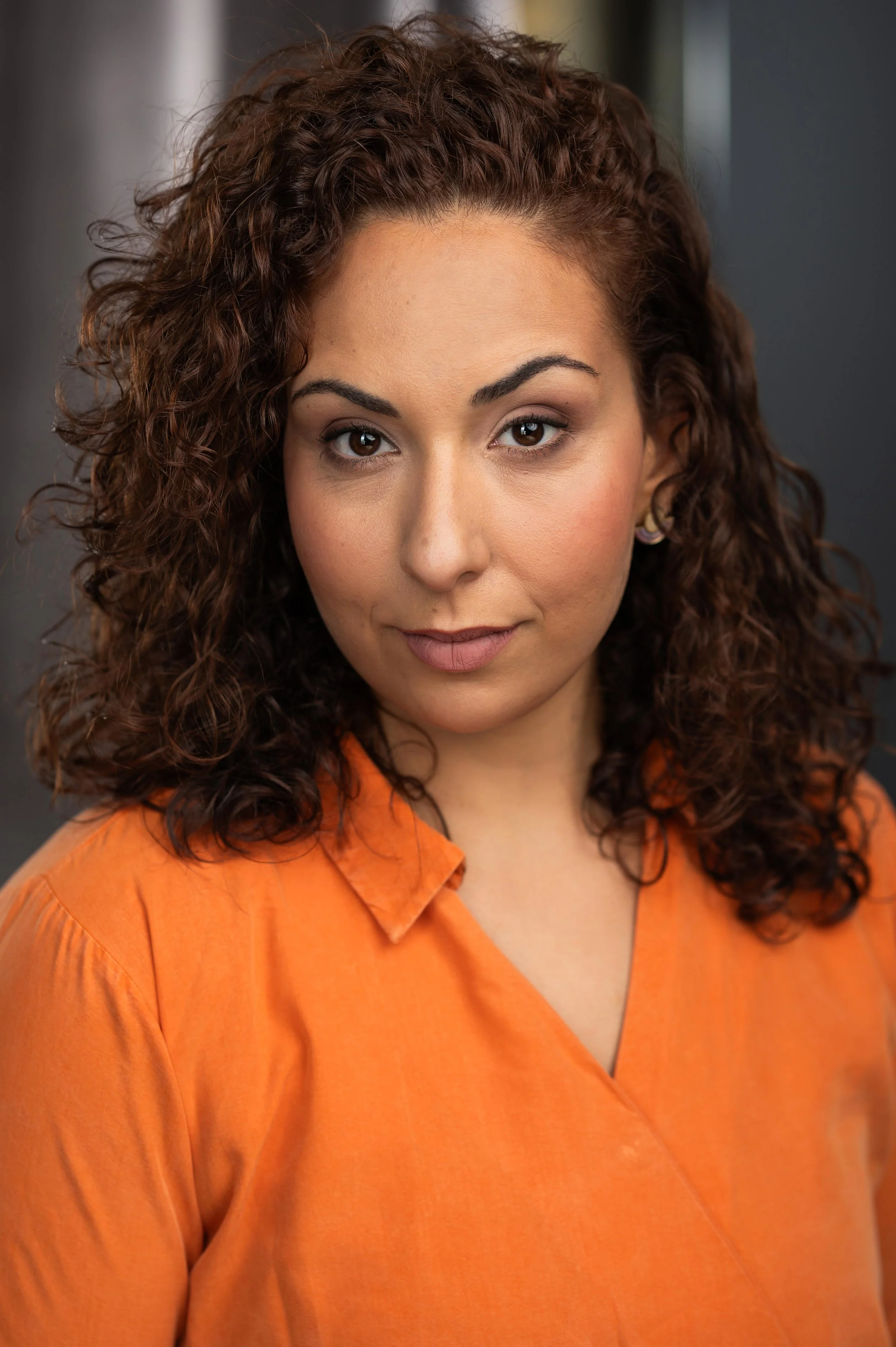 A woman with curly brown hair wearing an orange shirt, looking directly at the camera.