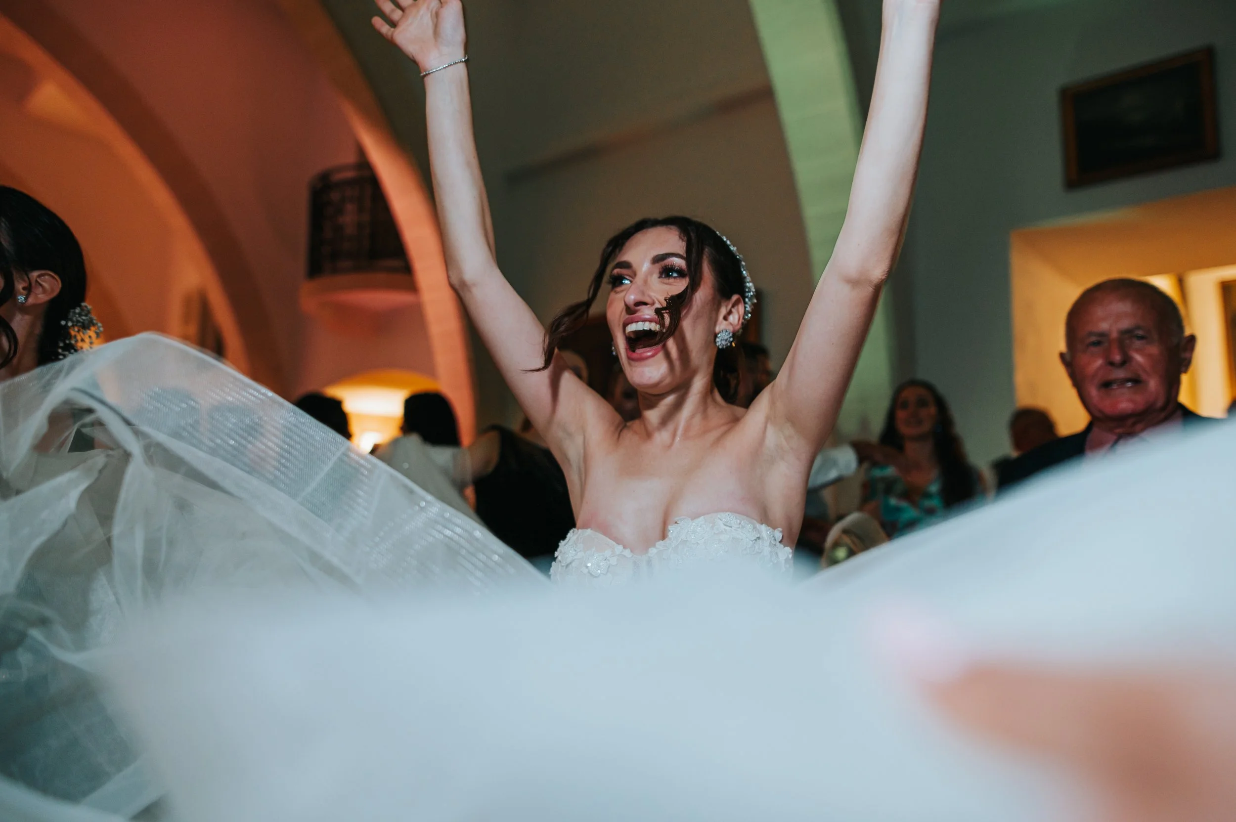 A joyful woman in a wedding dress celebrating with her arms raised at a wedding reception surrounded by guests.