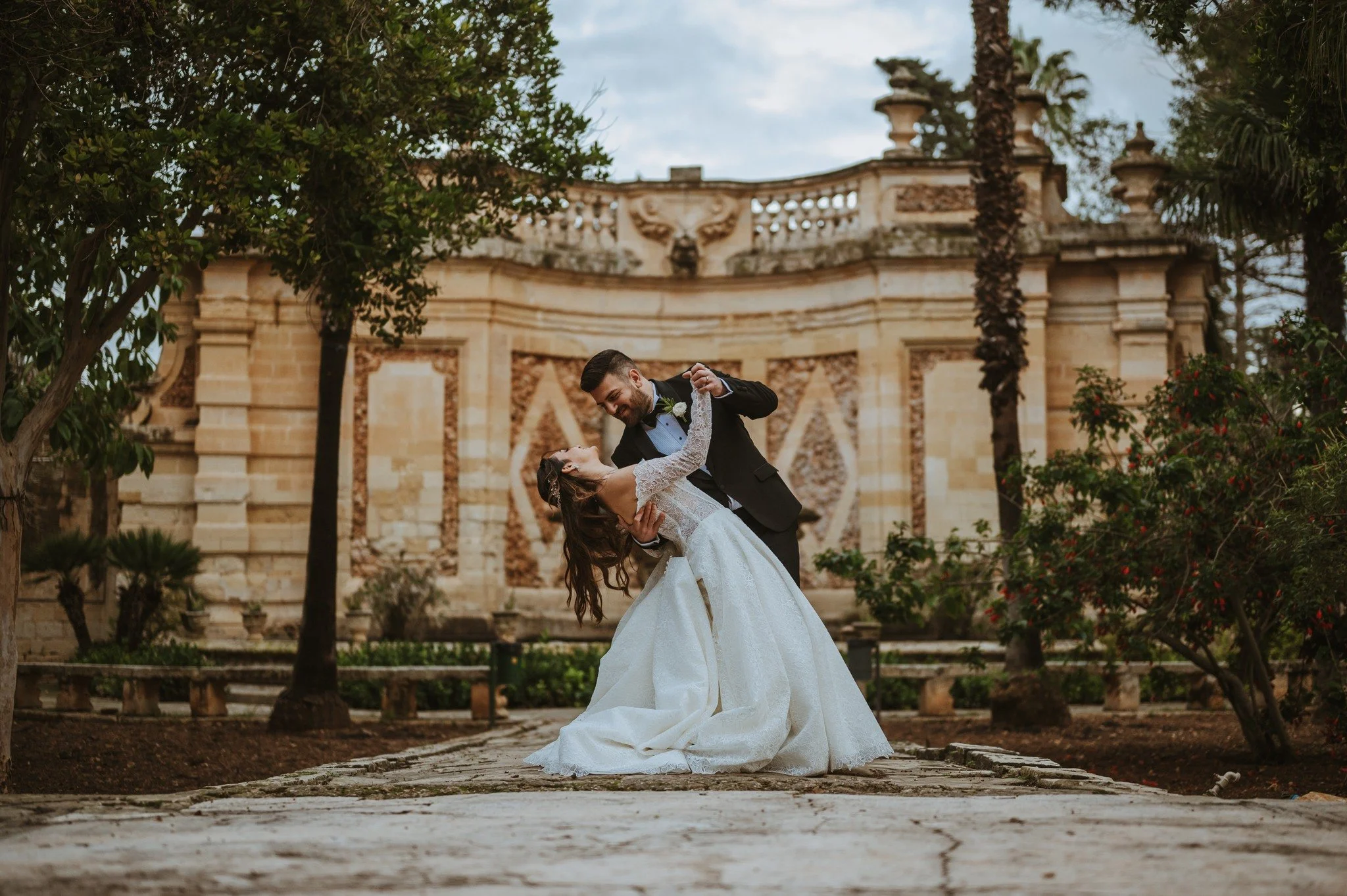A wedding couple dancing outdoors in front of a historic stone building with tall trees surrounding them.