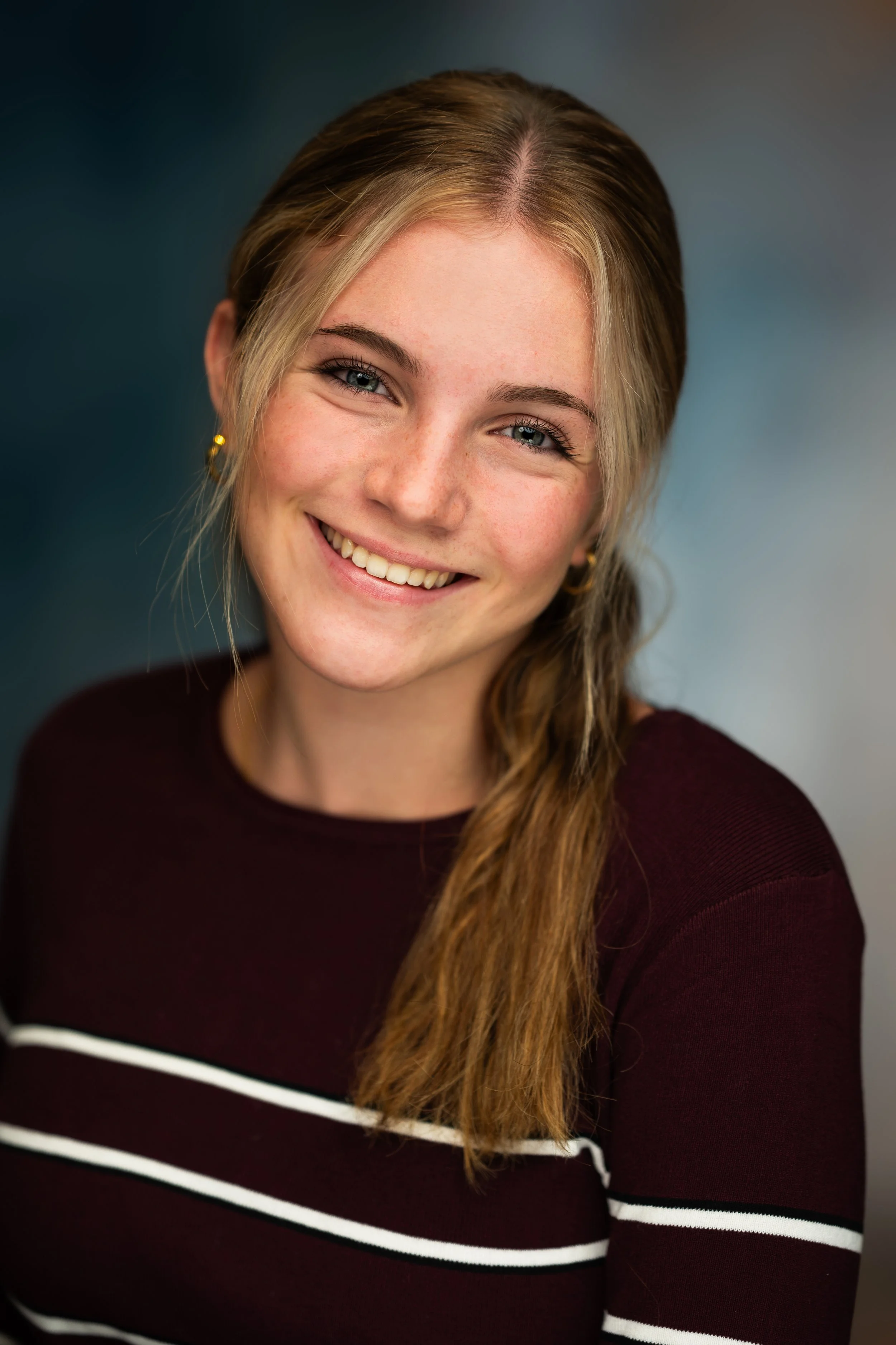 Close-up of a young woman with long, wavy light brown hair and blue eyes, smiling, wearing a maroon sweater with white and black stripes and small gold earrings.