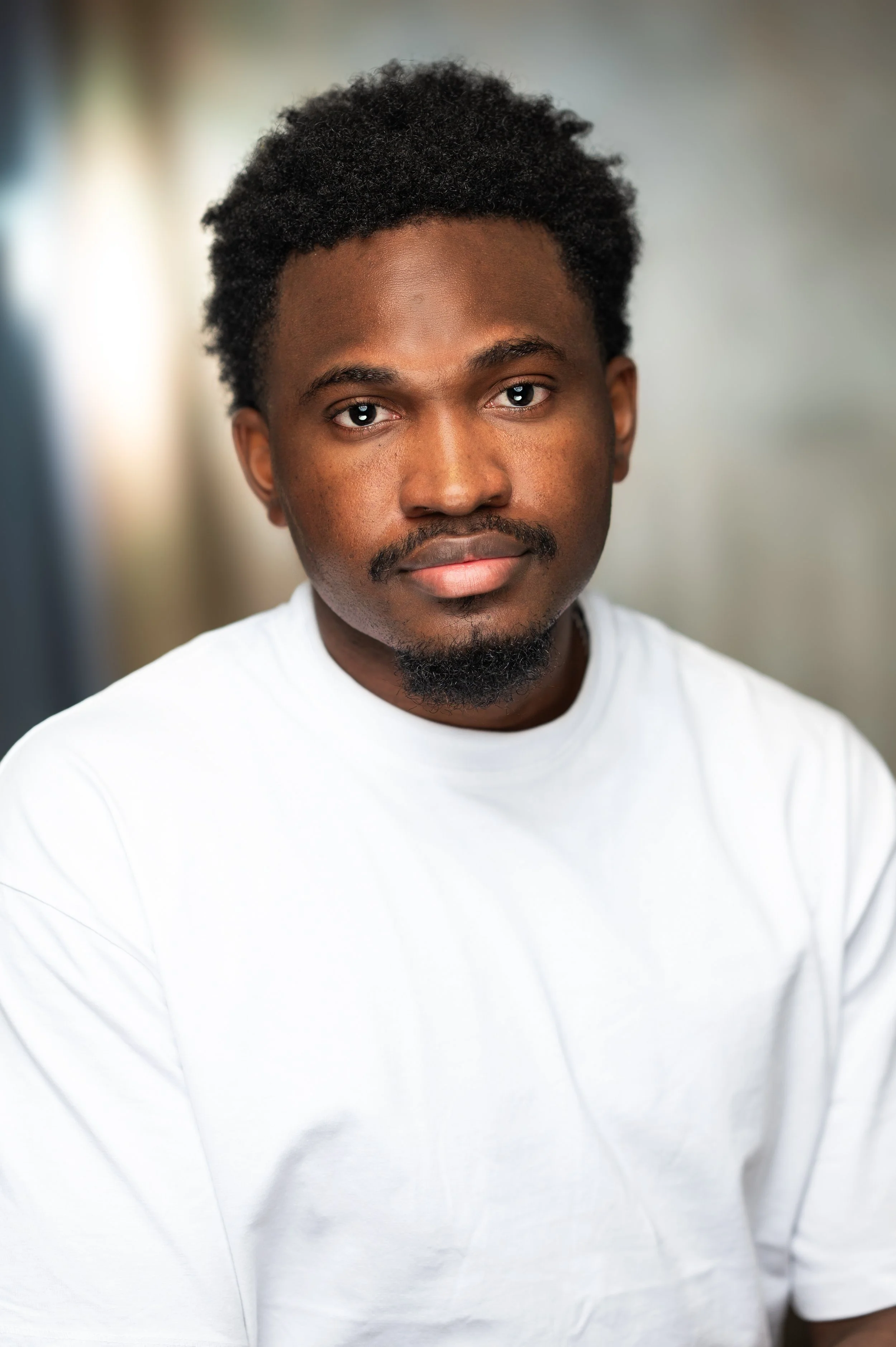Close-up portrait of an African American man with black curly hair, slight beard, wearing a plain white t-shirt, against a blurred background.