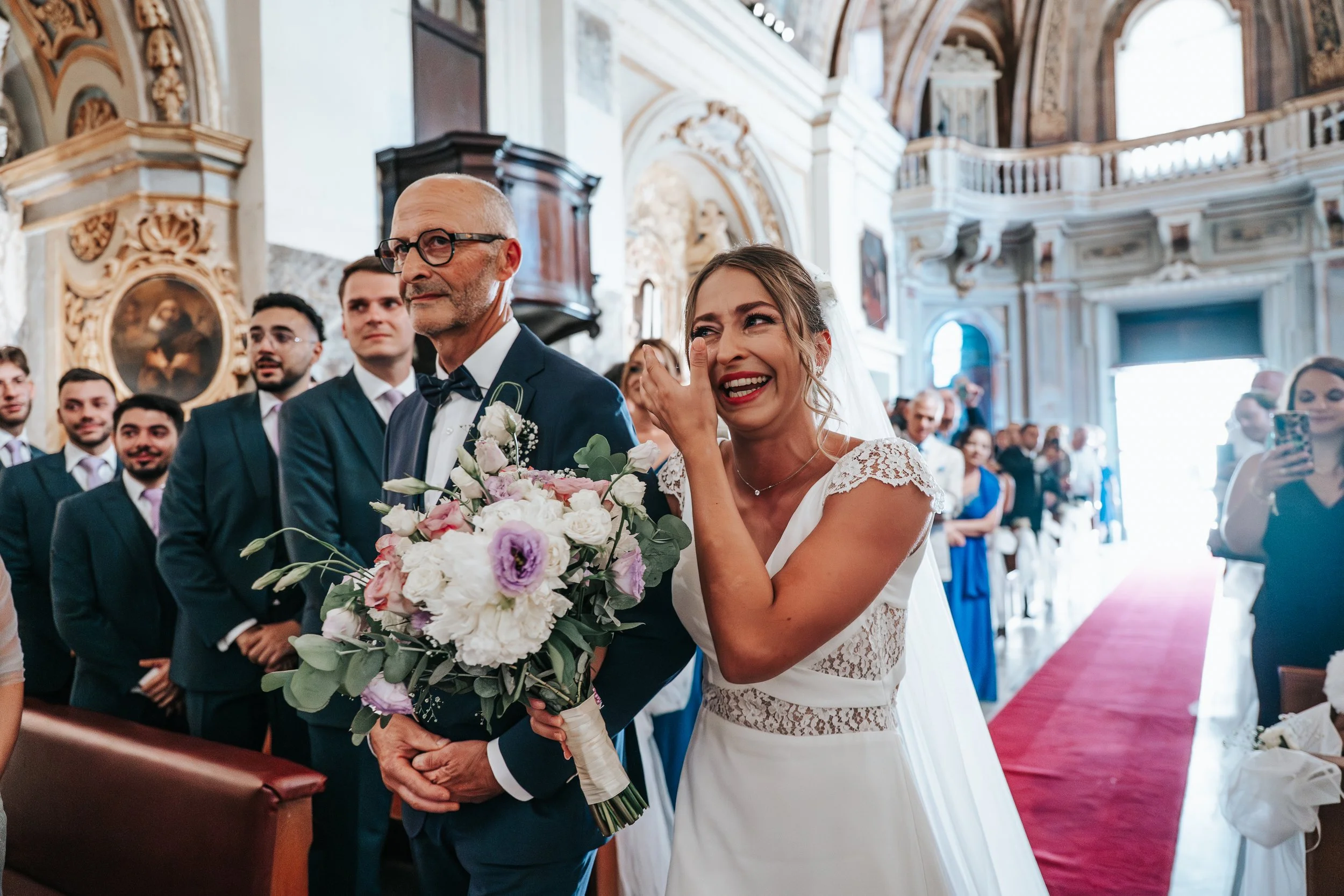 A bride is laughing and wiping tears in a church during her wedding ceremony, holding a bouquet of flowers, with guests seated behind her.