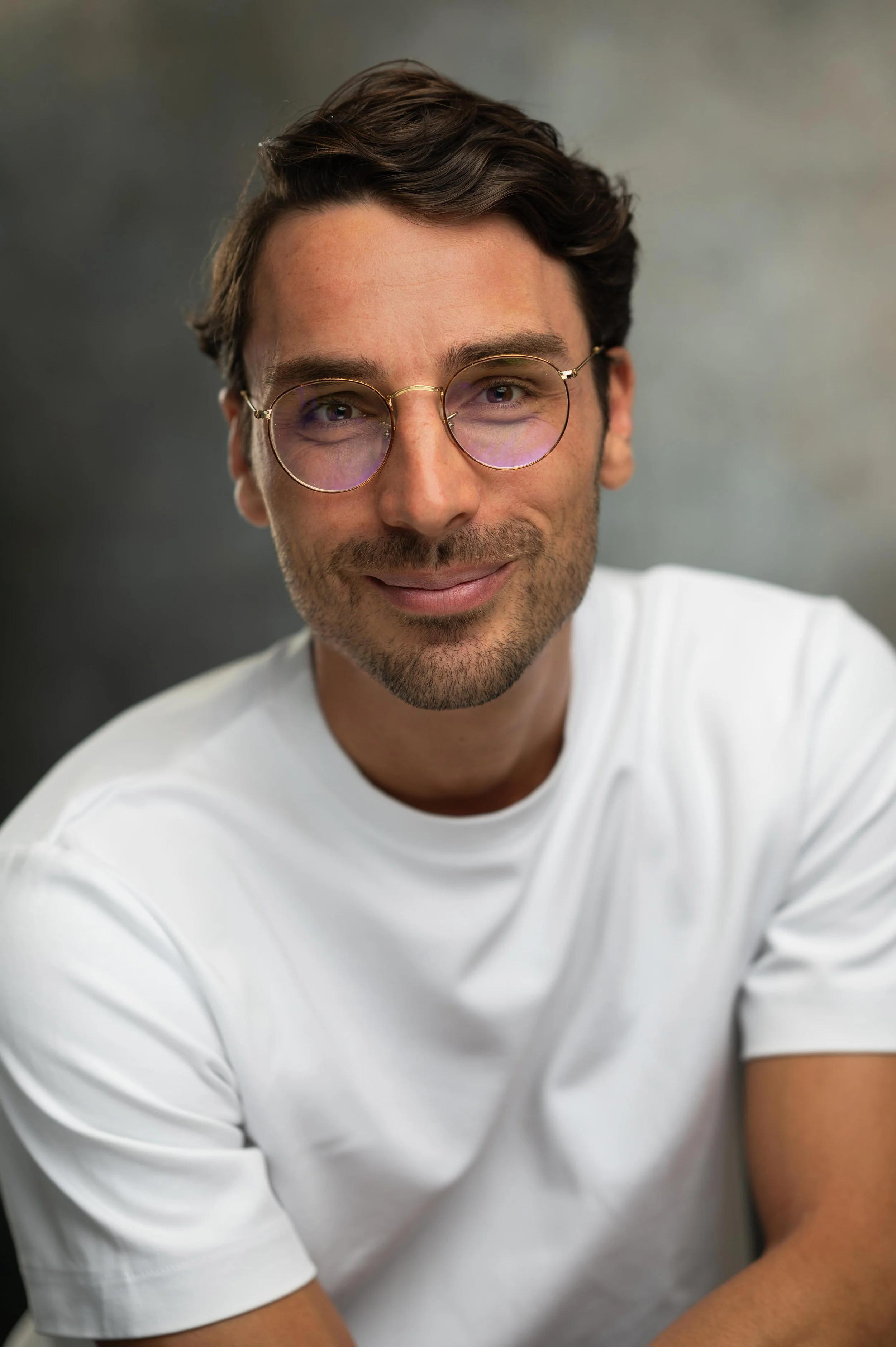 Close-up of a young man with dark wavy hair, wearing round glasses with a gold frame and a white t-shirt, smiling at the camera against a neutral background.