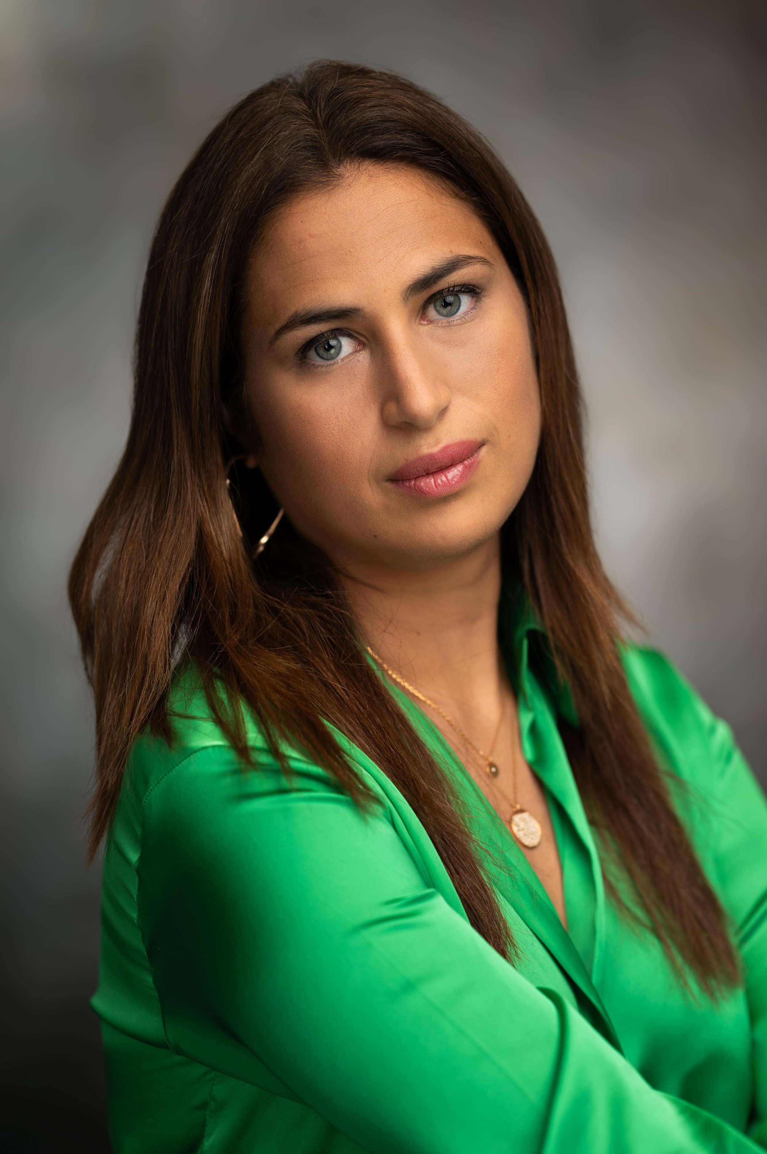 A woman with long brown hair wearing a bright green shirt and gold jewelry, looking into the camera with a neutral expression, against a blurred gray background.