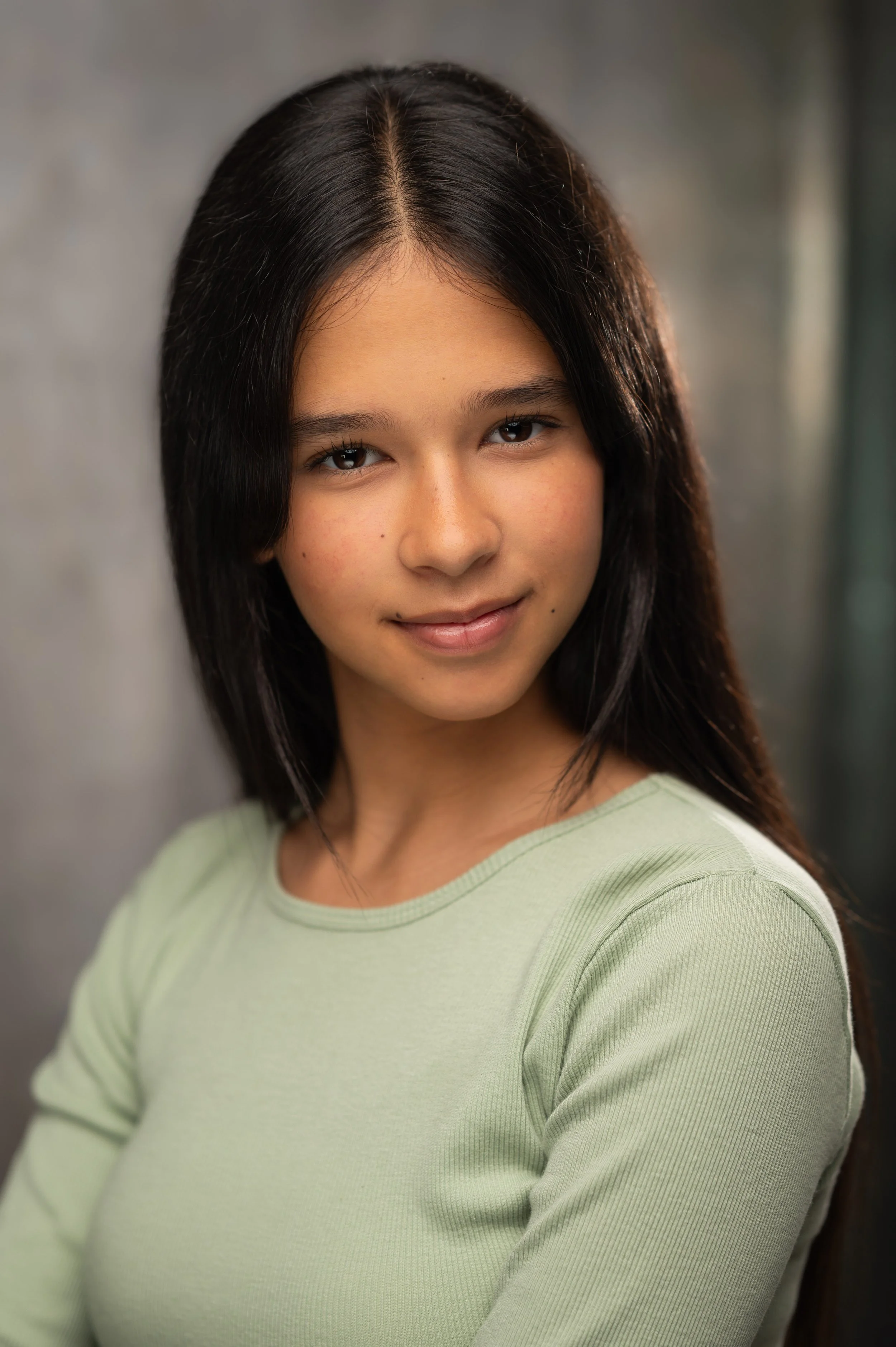 A young girl with long black hair and light skin, wearing a light green shirt, standing against a blurred background.