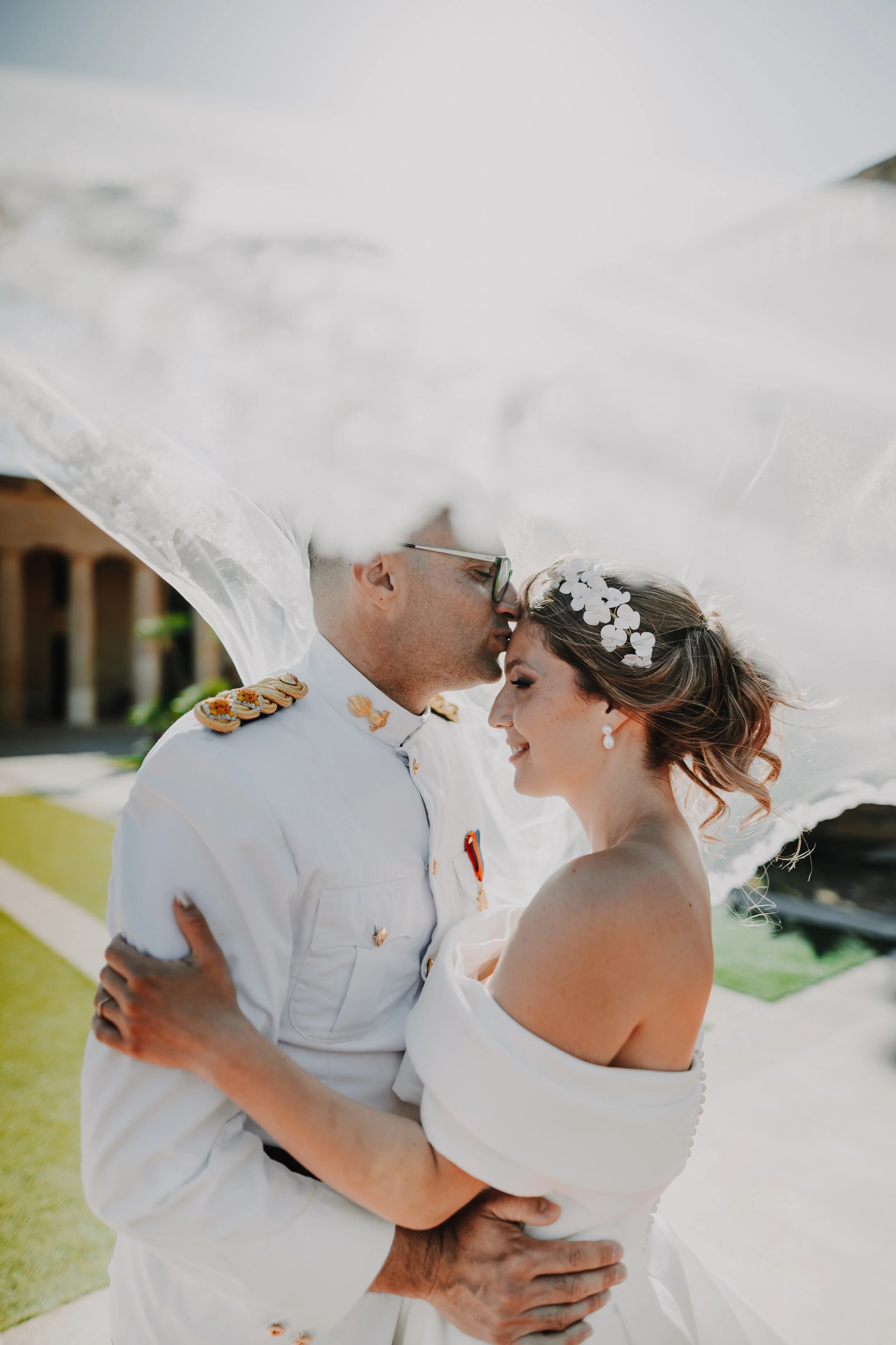 A couple dressed in white, sharing a kiss while dancing outdoors, with a background of water and a dock.