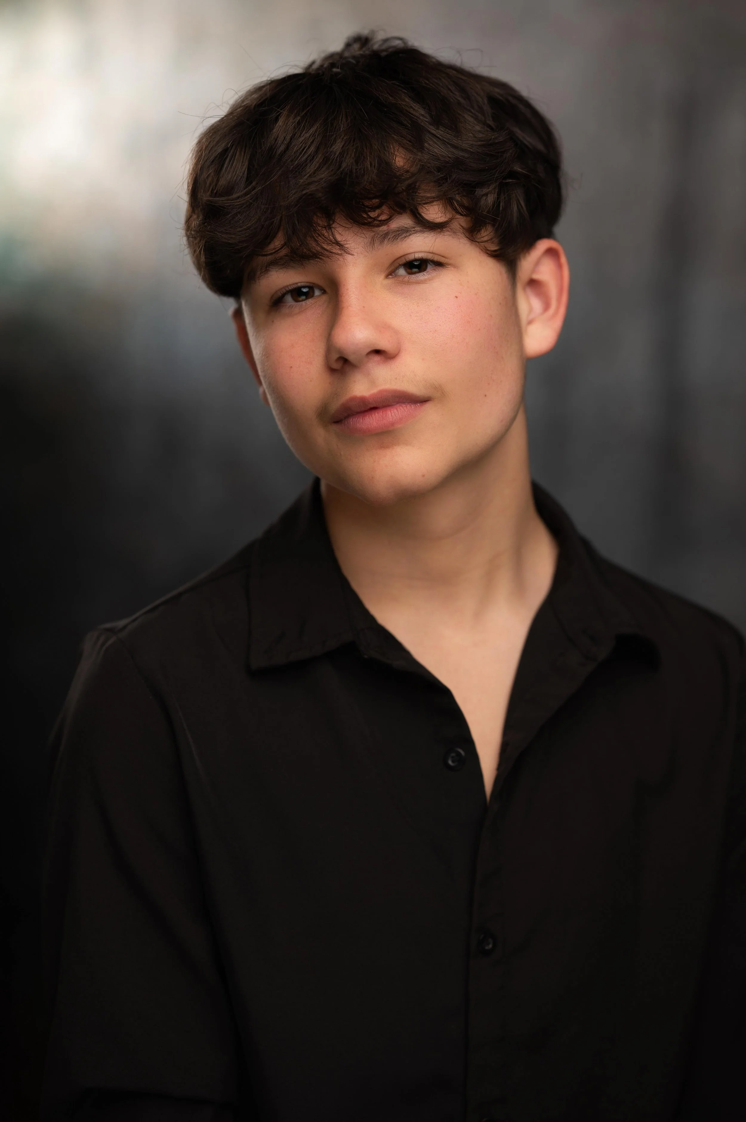 A young person with dark brown hair looking at the camera, wearing a black button-up shirt, against a dark gray blurred background.