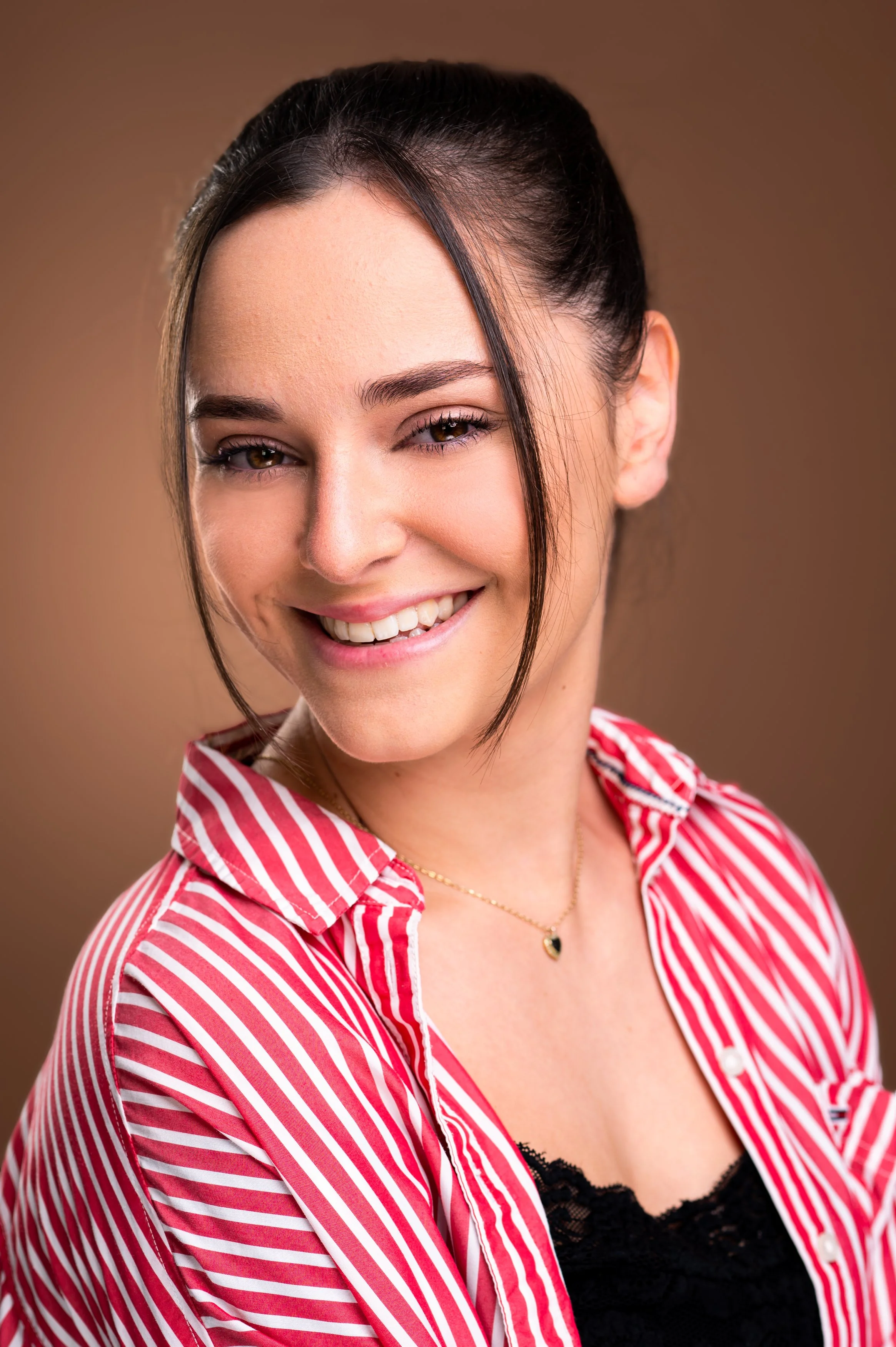 A young woman with dark hair styled in an updo, smiling, wearing a red and white striped shirt and a necklace with a heart pendant, against a plain brown background.